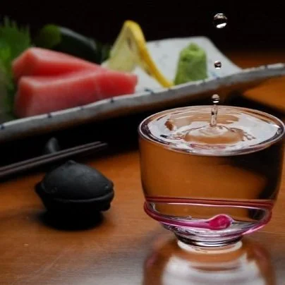 A glass cup of tea with a drop falling into it, placed on a wooden table, with sushi, wasabi, and ginger in the background.