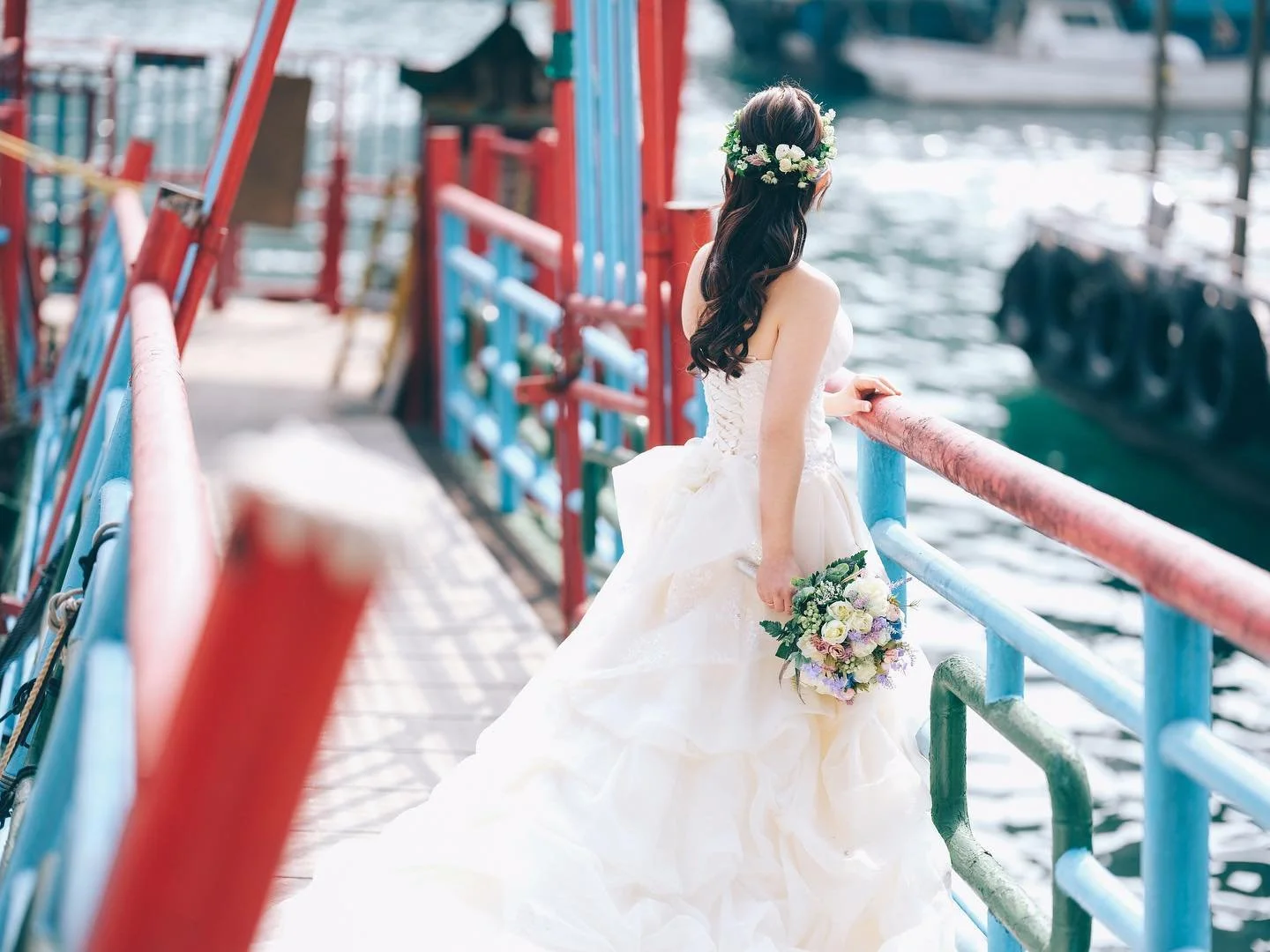Bride in a strapless white wedding dress with long dark hair and a floral crown, holding a bouquet, standing on a colorful dock by the water, looking away from the camera.