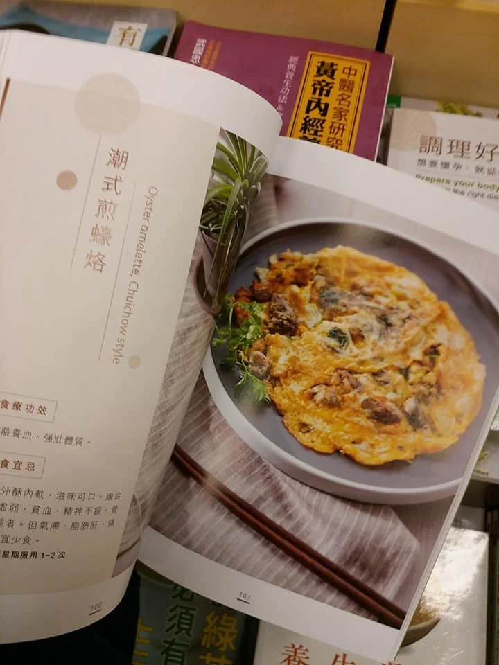 A page from a cookbook featuring an oyster omelette with chives, served on a gray plate with chopsticks and a sprig of herbs for garnish.