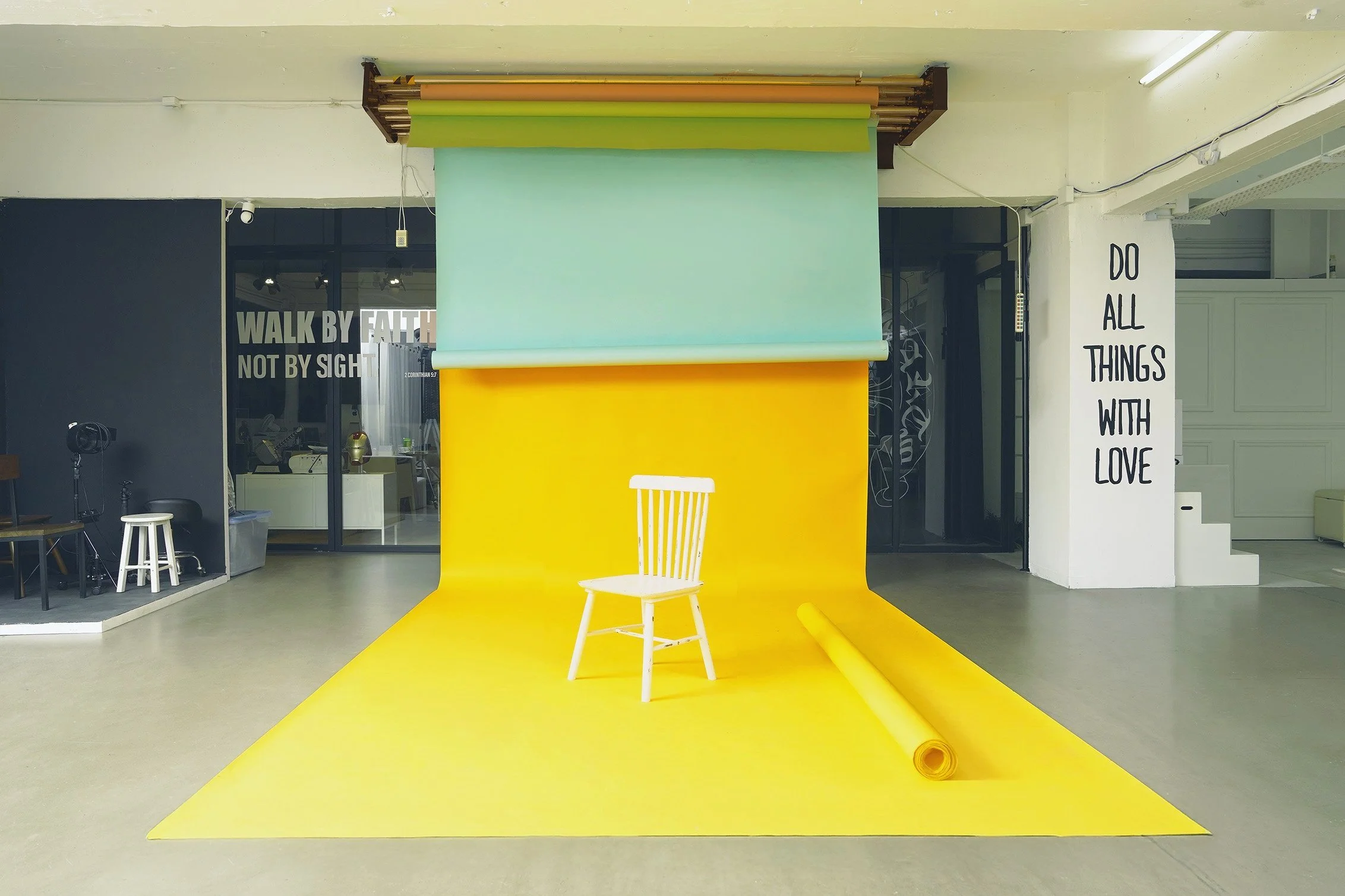 Colorful photography studio with yellow backdrops, a white chair, and a roll of yellow paper, with inspirational wall texts 'WALK BY FAITH, NOT BY SIGHT' and 'DO ALL THINGS WITH LOVE'.
