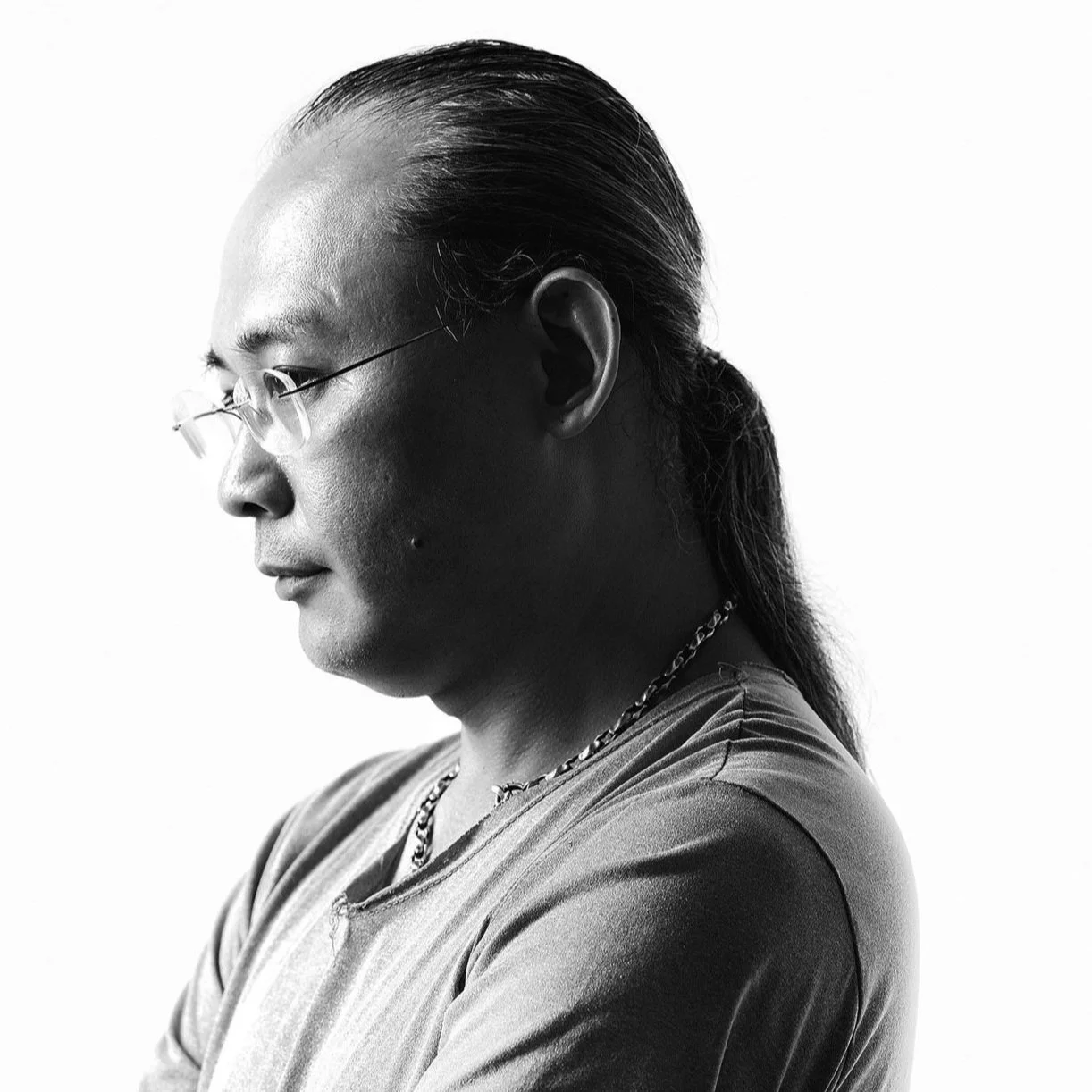 Black and white side profile of a man with long hair tied back, wearing glasses, a chain necklace, and a t-shirt.