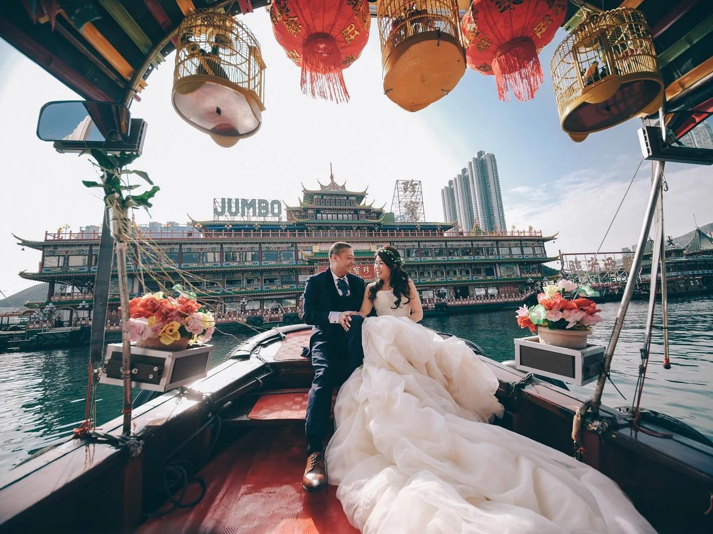 A wedding couple sitting on a boat with a Chinese temple-style building and city skyline in the background, decorated with lanterns and flowers.
