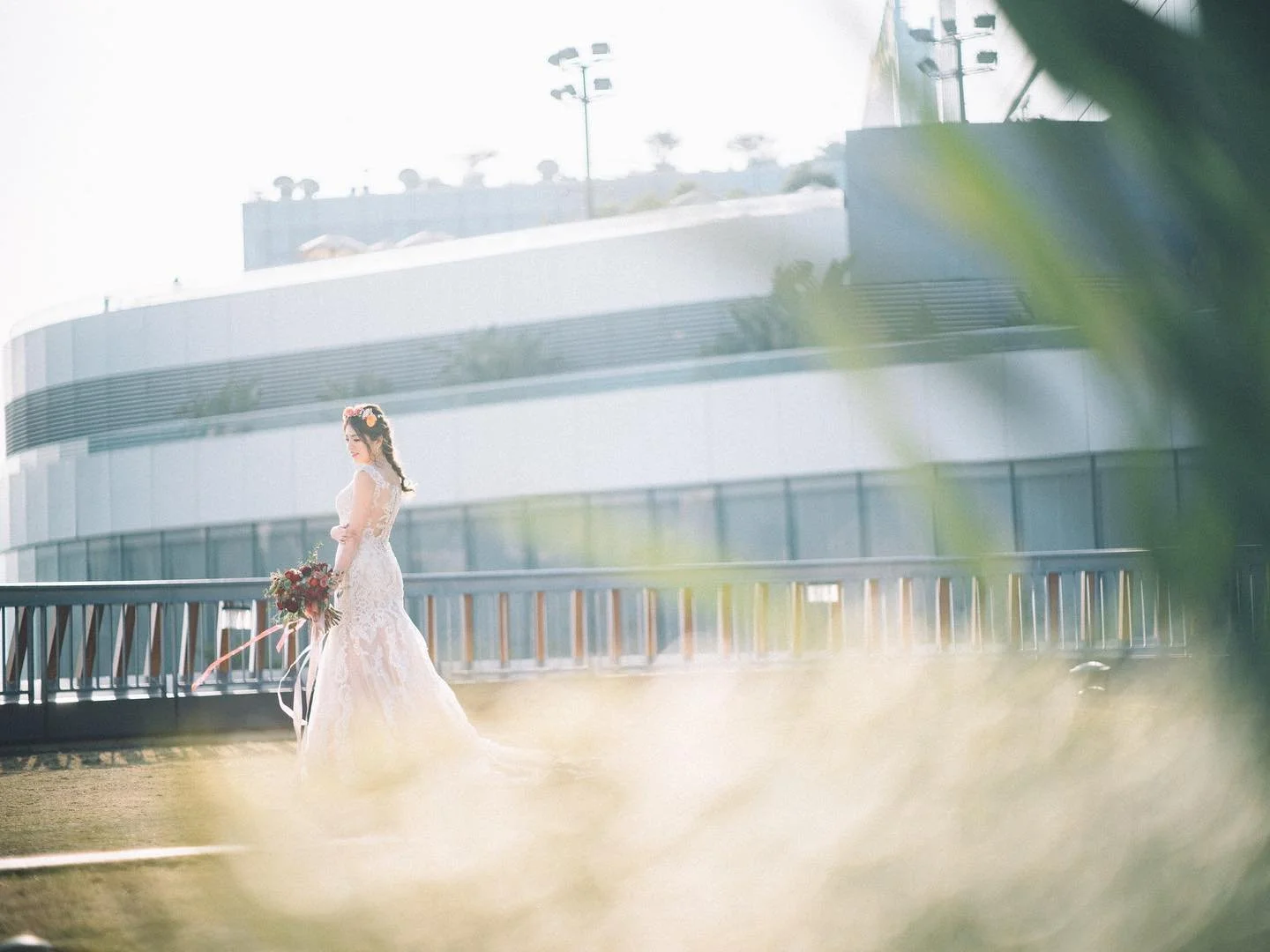 A woman in a wedding dress holding a bouquet, standing outside on a balcony or terrace in daylight, with an abstract modern building in the background and greenery partially obscuring the view.
