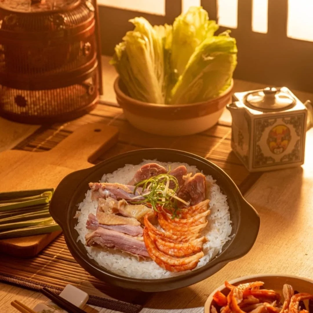 A bowl of rice topped with sliced meat, shrimp, and garnished with green onion, on a wooden table with side dishes, a bowl of lettuce, a teapot, and chopsticks.