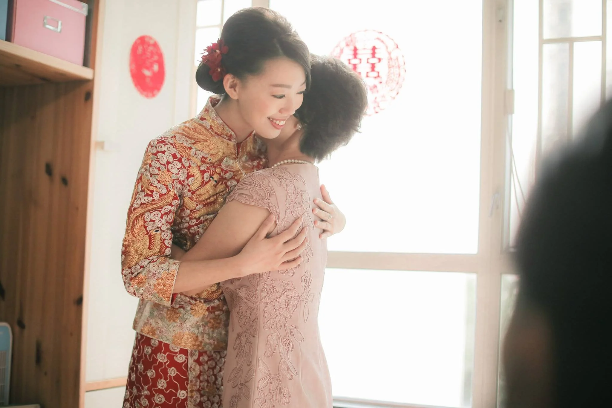 A woman in a traditional Chinese dress hugging another woman in a pink dress near a window with red decorations.