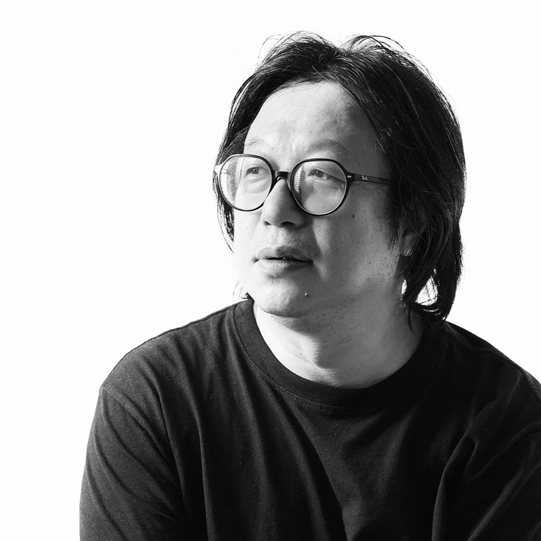 Black and white portrait of an Asian man with medium-length hair and glasses, looking to the side, wearing a dark T-shirt.