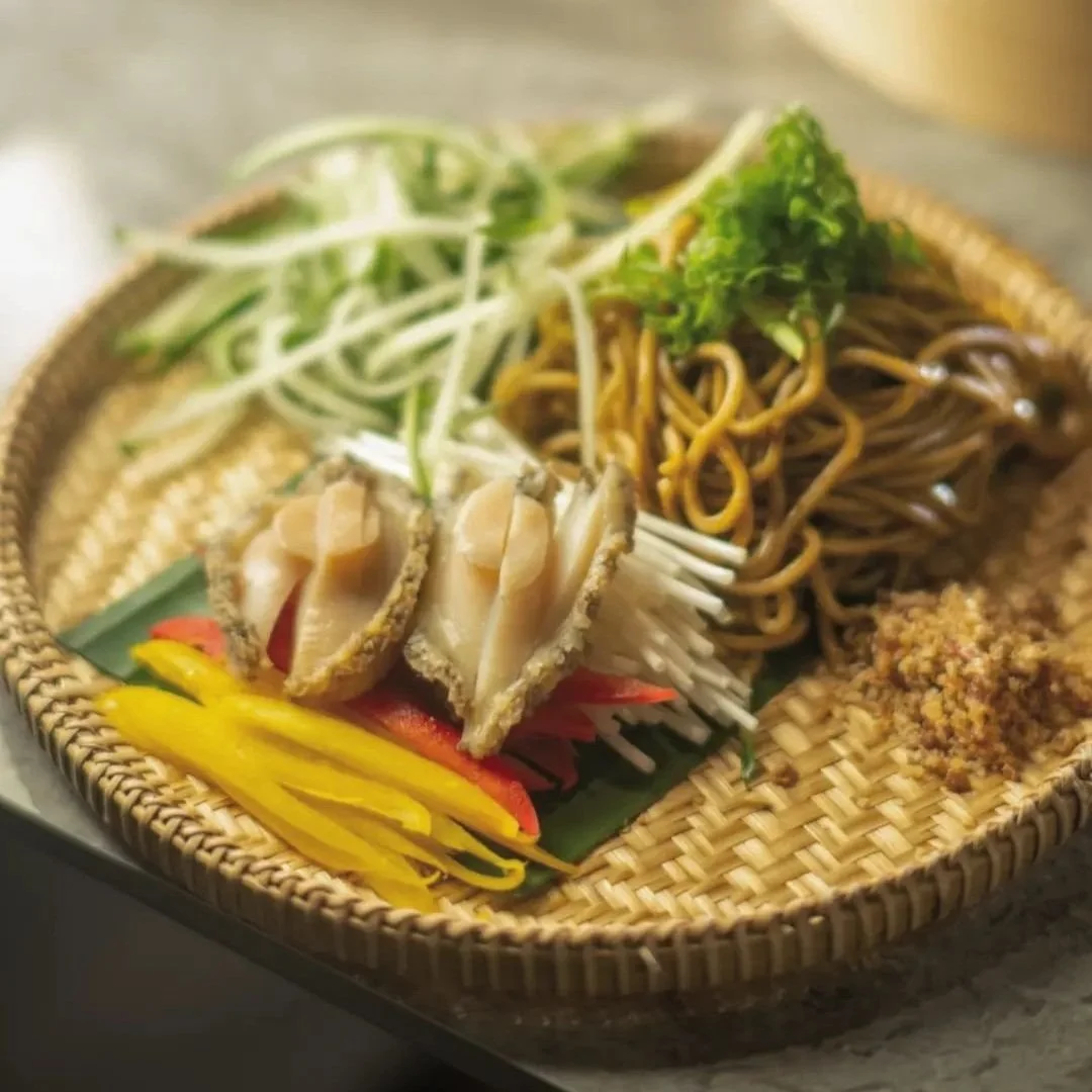 Assorted Asian cold noodle dish with vegetables, sliced chicken, and garnishes on a woven bamboo platter.