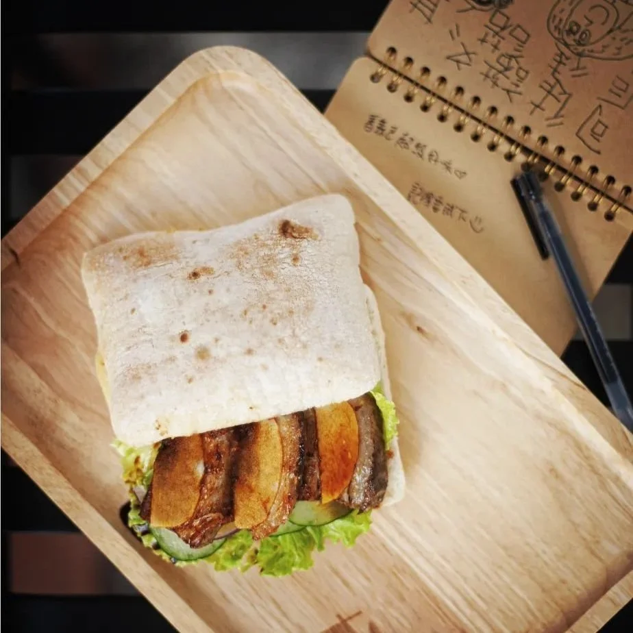 A sandwich with bacon, sliced cucumber, lettuce, and flatbread on a wooden tray, with a notebook and pen nearby.