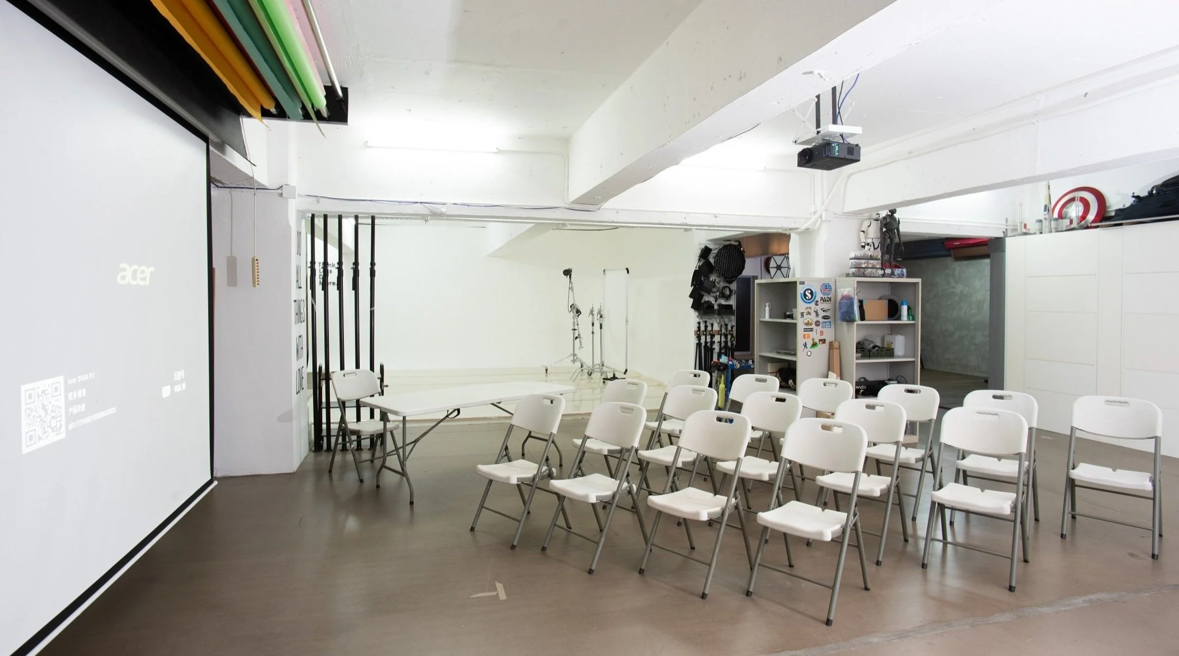Room with white chairs set up in rows for a presentation or meeting, a projector mounted on the ceiling, a screen displaying an Acer logo, and equipment including lighting and storage shelves in the background.
