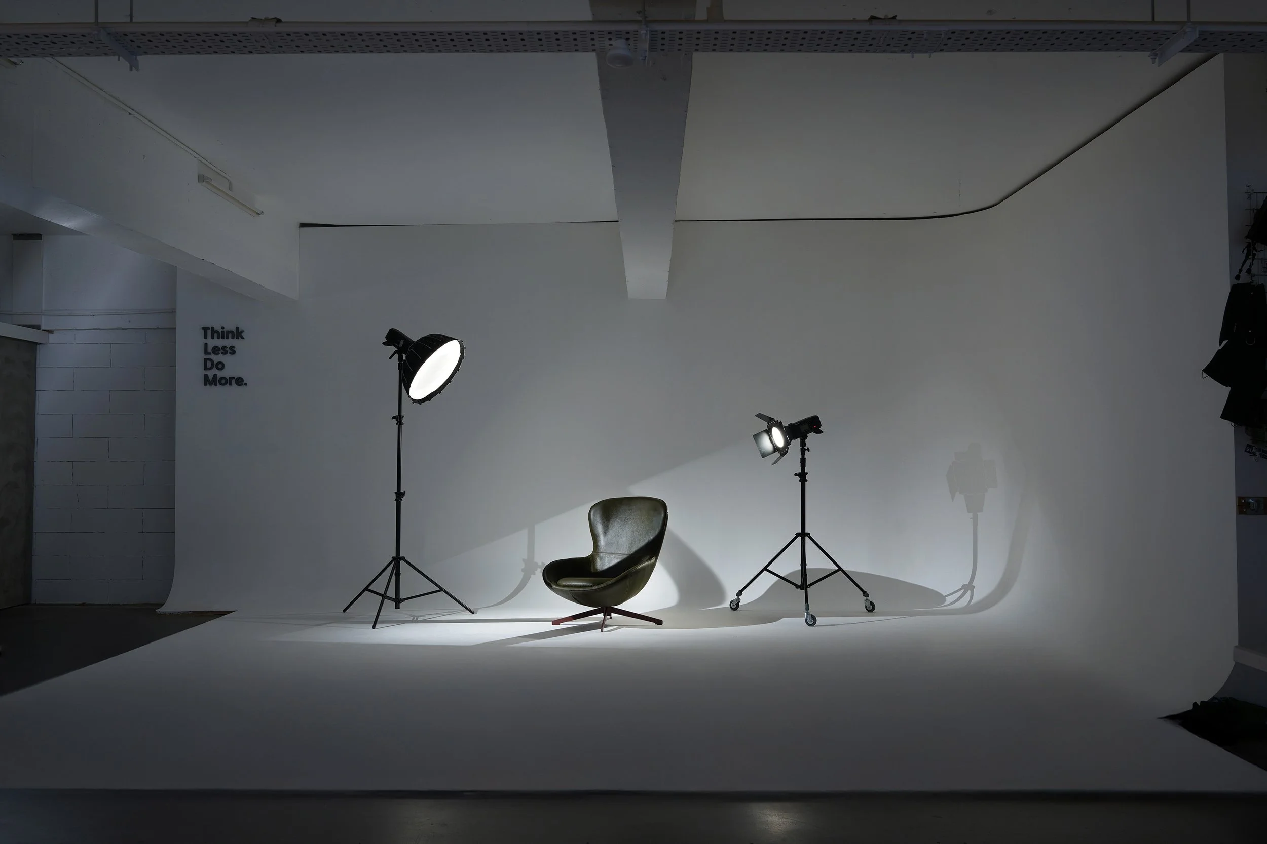 Photography studio setup with a black leather chair, two studio lights on tripods, and a white backdrop, with the words 'Think Less Do More' on the wall.