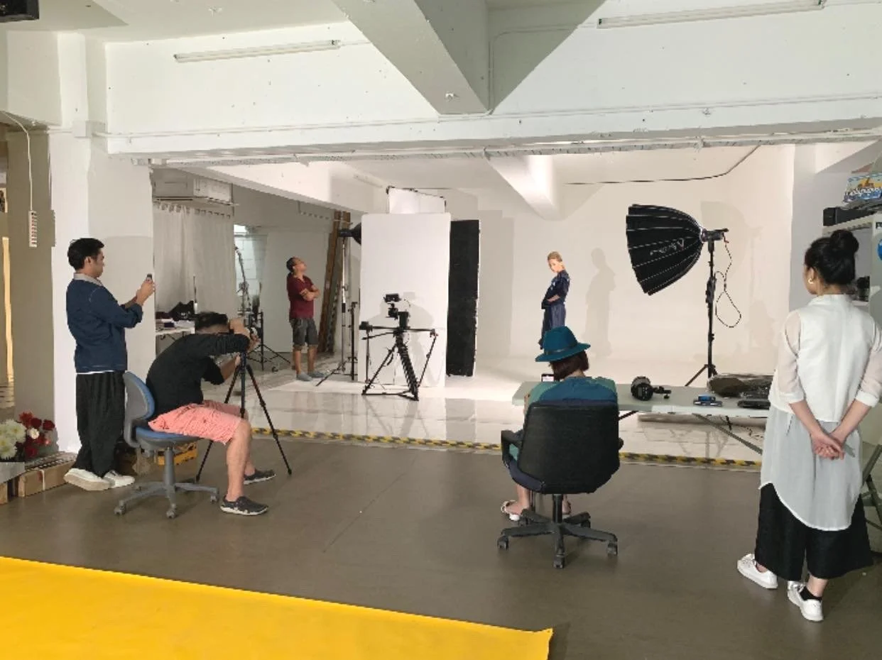 Indoor photography studio with a model standing in front of a white backdrop, surrounded by crew members and equipment including cameras, lights, and tripods.