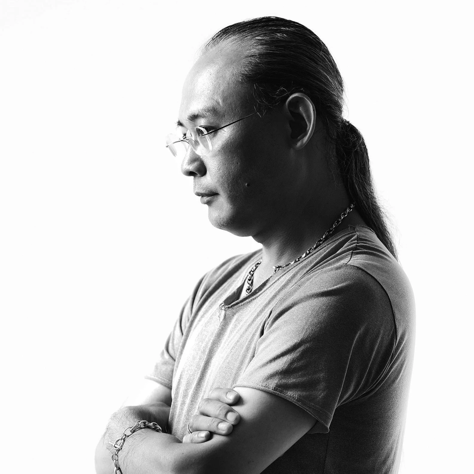 Black and white portrait of a man with long hair tied back, wearing glasses, a chain necklace, a bracelet, and a t-shirt, standing with arms crossed against a plain background.