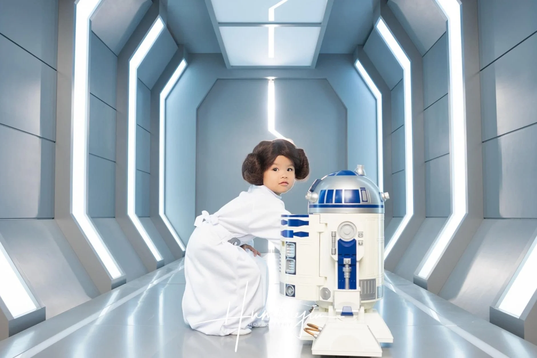 A young girl dressed as Princess Leia from Star Wars in a sci-fi corridor with LED lights, kneeling next to a replica of R2-D2 robot.