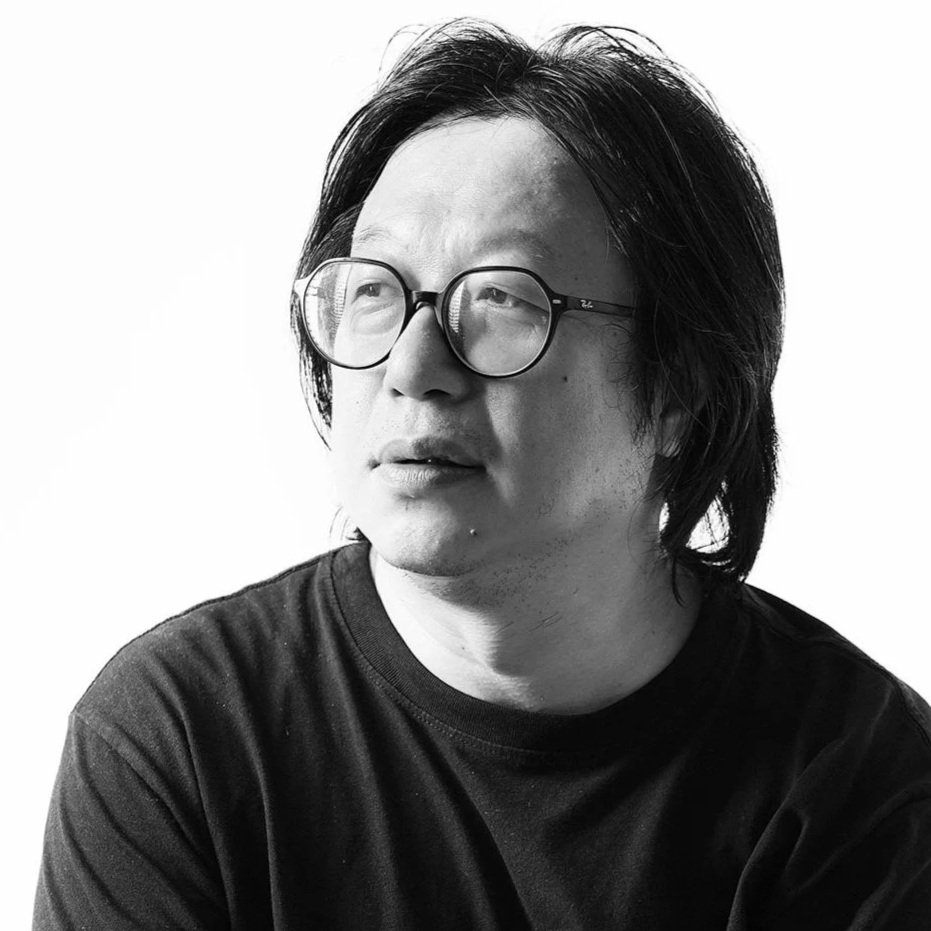 A black-and-white portrait of a man with medium-length dark hair and glasses, wearing a dark shirt, looking slightly to the side against a plain white background.