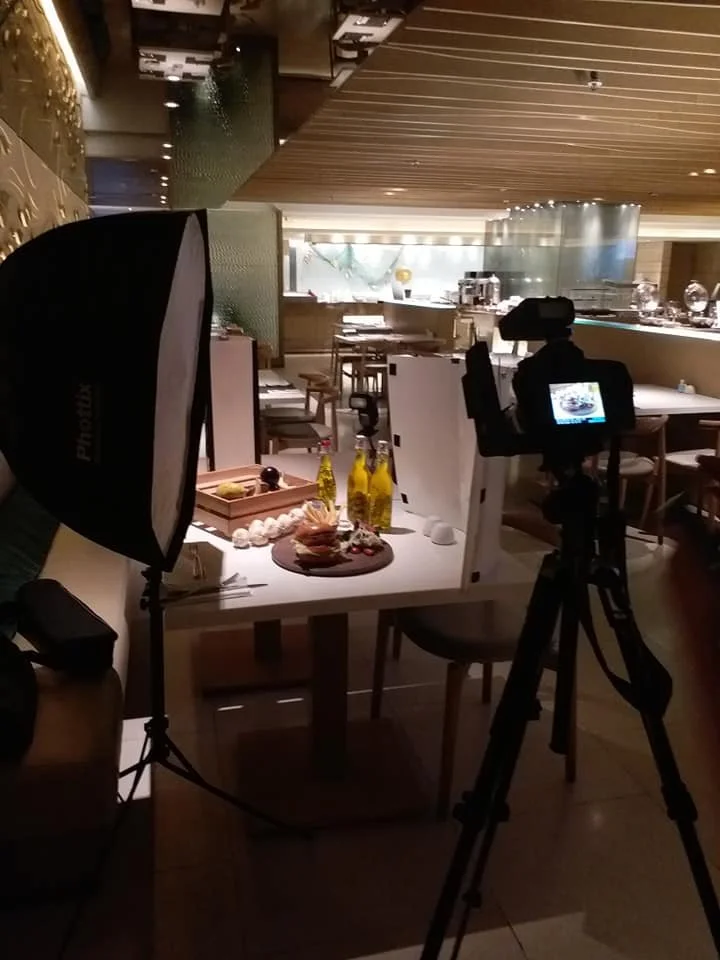 Photography setup in a restaurant or cafe with a softbox light on the left, a camera on a tripod facing a table with bottles and food items, and a background of a well-lit, modern interior with dining tables and a bar area.