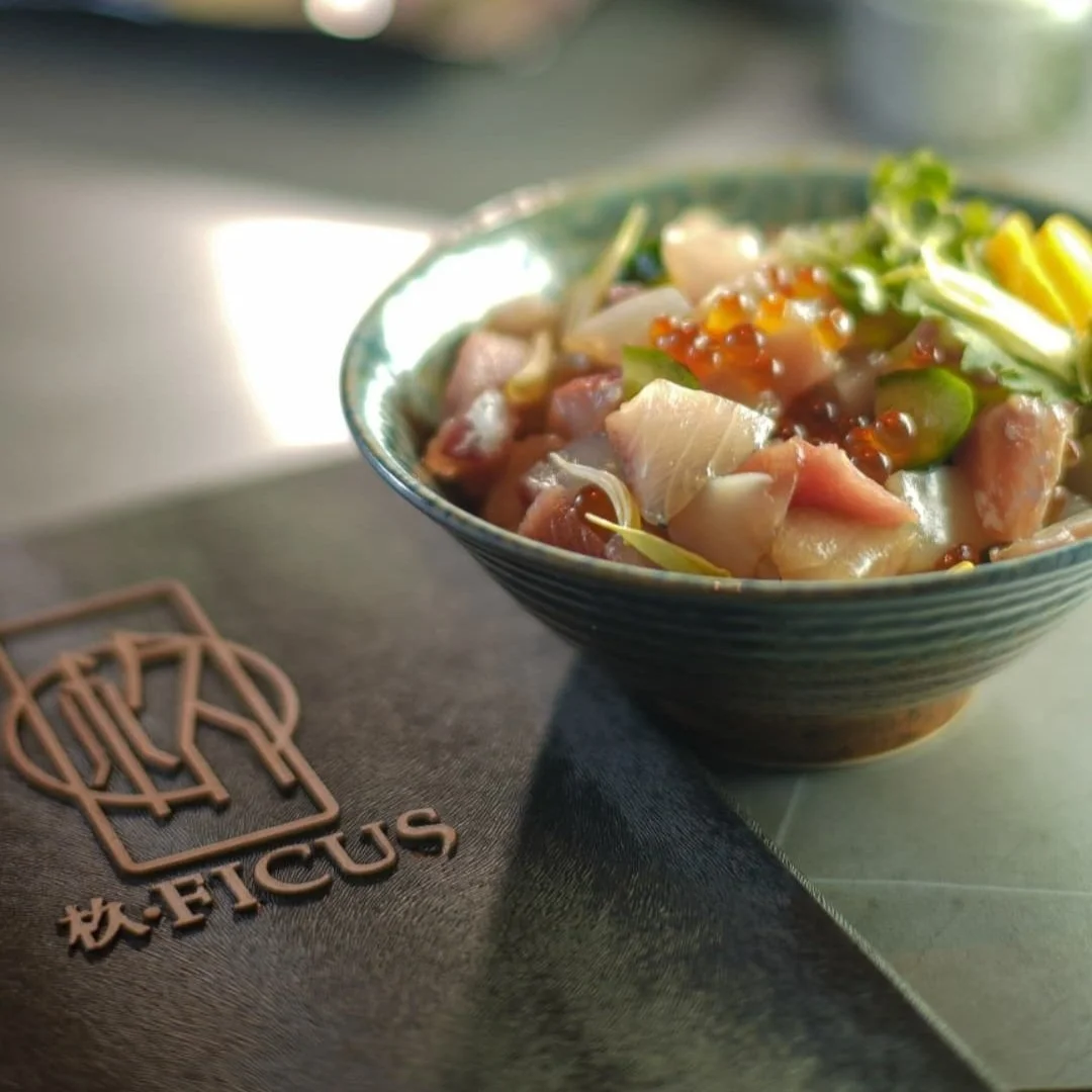 Close-up of a bowl of poke salad with raw fish, vegetables, and seasonings on a table with a Ficus logo on a cloth.
