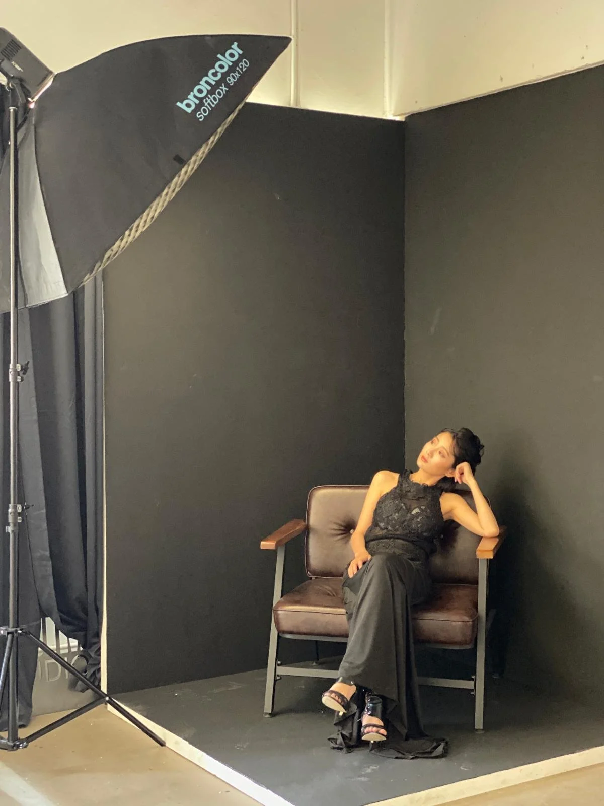 A woman in a black lace top and wide-leg black pants sitting on a vintage-style brown leather and wood armchair in a photography studio with black walls and a large softbox light.