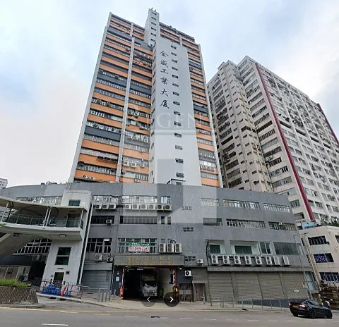 Tall multi-story building with orange and gray exterior, featuring Chinese characters on its facade, situated among other high-rise buildings, with street-level shops and parked cars in front.