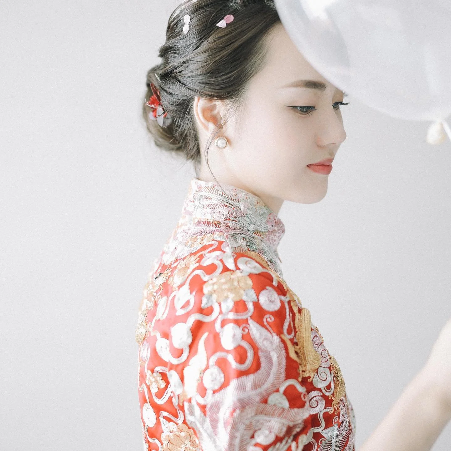 A woman dressed in a traditional red and gold Chinese wedding dress with intricate embroidery, looking down with a gentle smile.