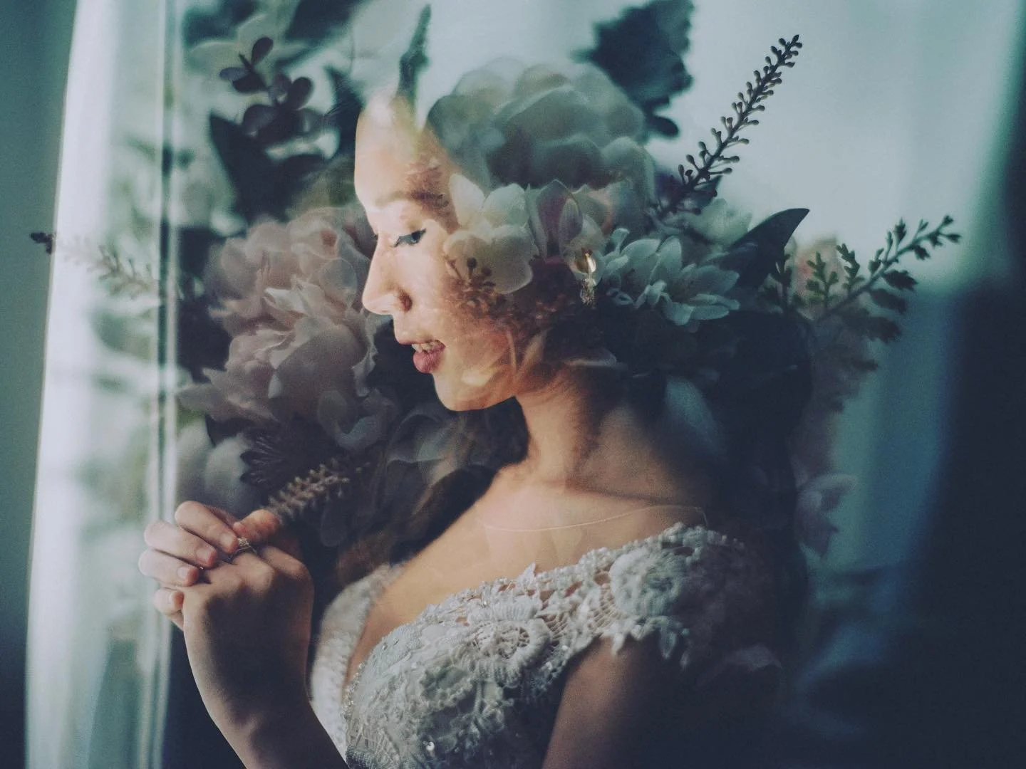 A woman with a floral headpiece, holding a feather, dressed in a lace gown, standing near a window with her eyes closed.