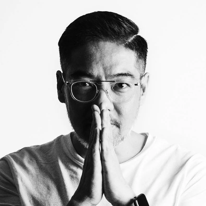 Black-and-white portrait of a man with glasses, short dark hair, and a beard, holding hands together in a prayer or meditation pose, facing the camera with a serious expression.