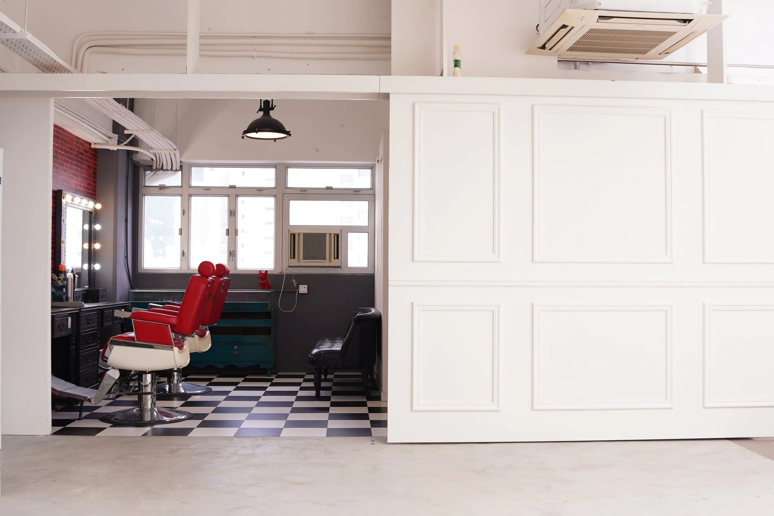 Interior view of a modern hair salon or barbershop with a red barber chair, a black chair, a large mirror, and a checkered black-and-white floor, with windows and an air conditioning unit.