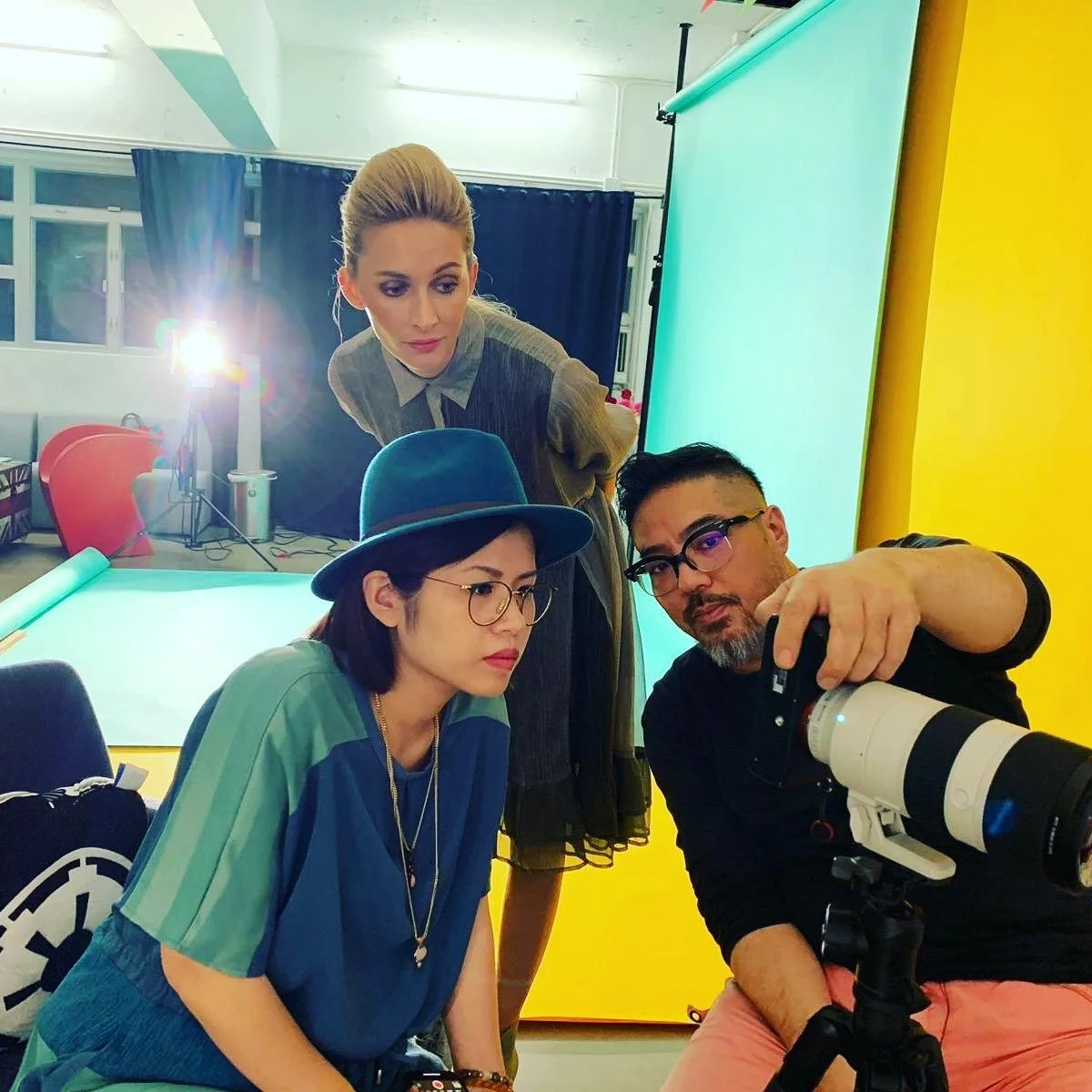 Three individuals, two women and one man, are in a photography or filming studio. They appear to be reviewing a shot, with one woman sitting, the other woman leaning over, and the man holding a camera with a large telephoto lens. The background includes a green screen and filming equipment.