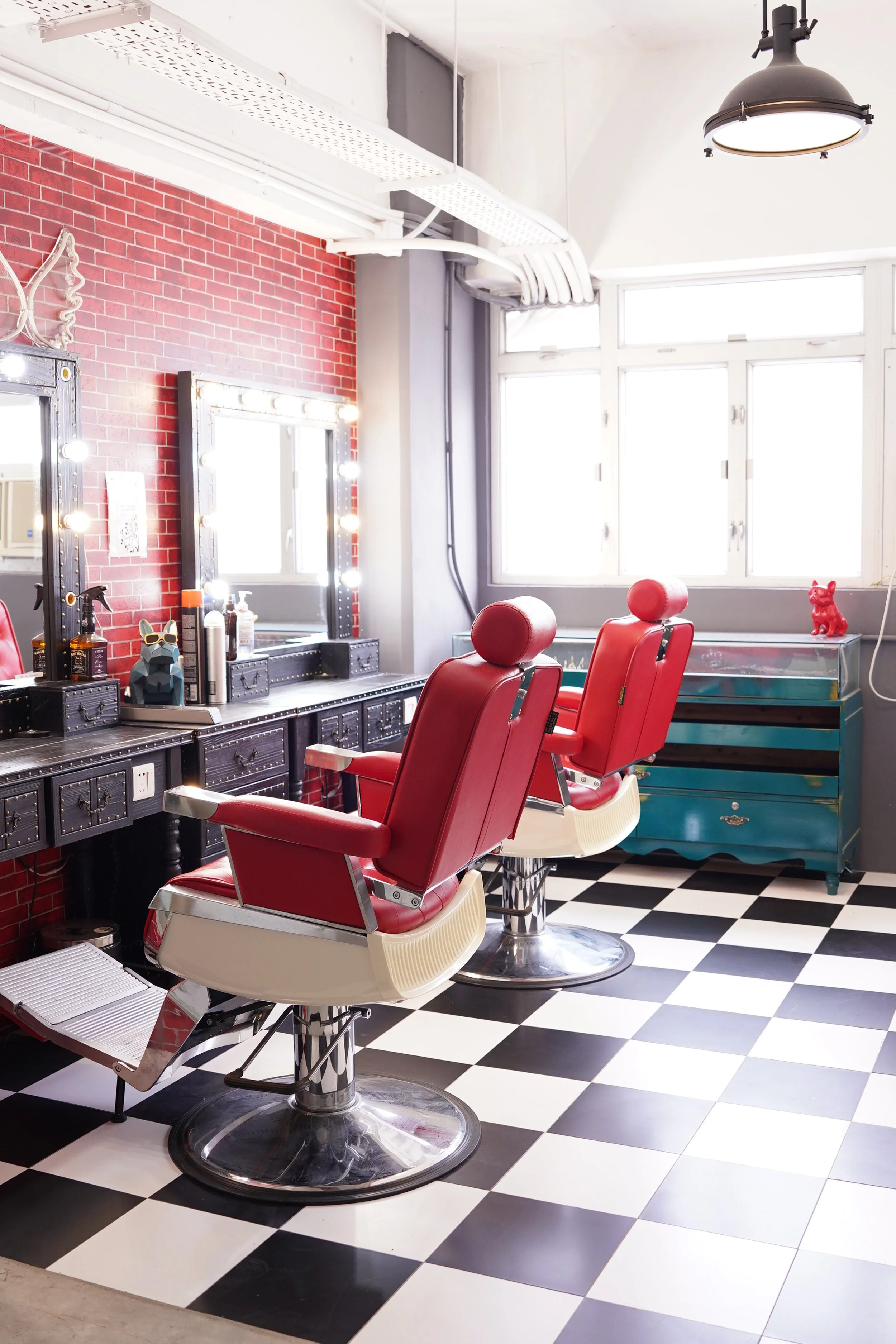 Interior of a vintage-style barbershop with two red barber chairs, black vanity mirrors with lights, and a window with natural light. The floor has a black and white checkered pattern, and there is a teal dresser against the wall.