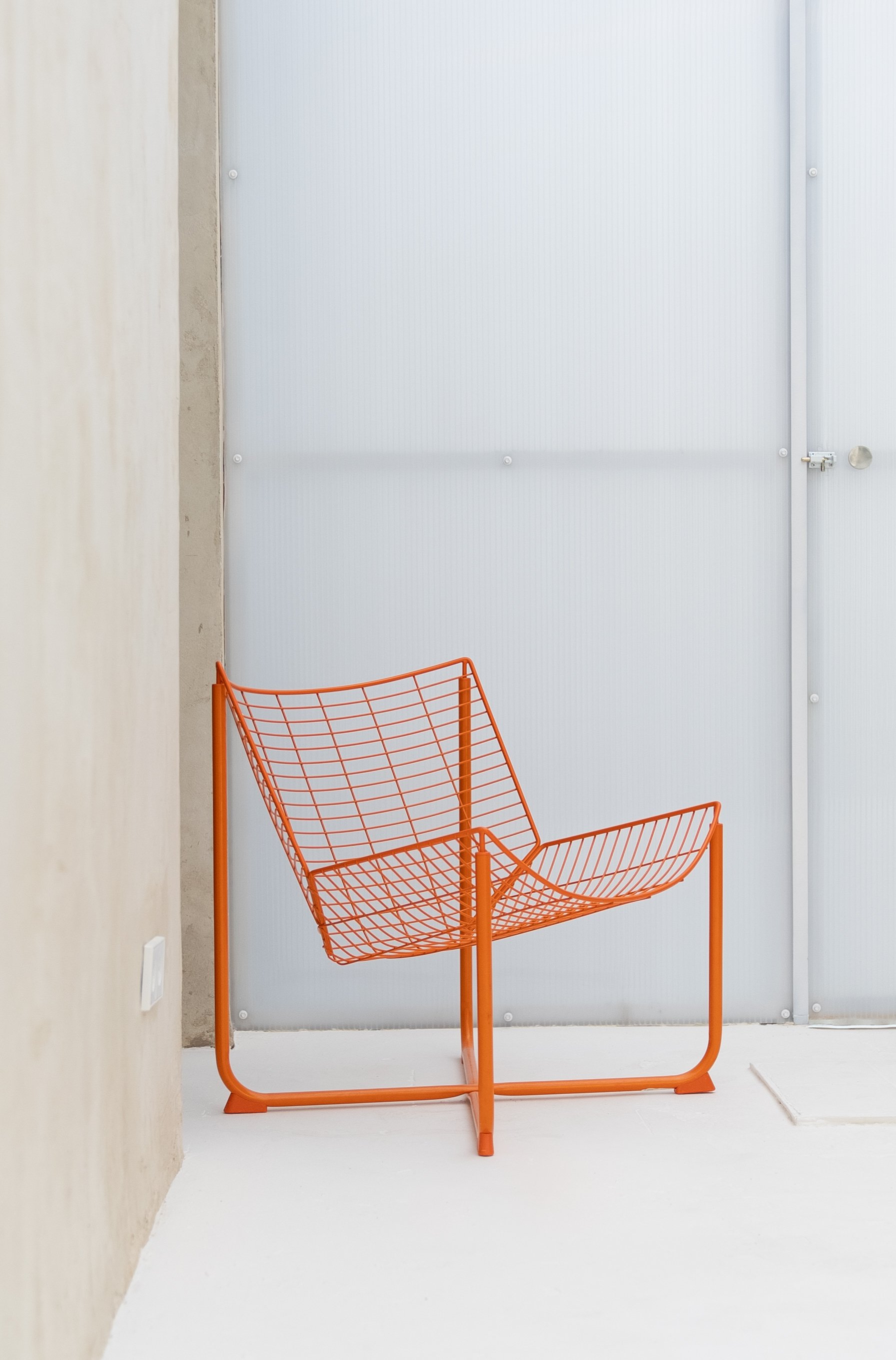 Orange wireframe chair in a minimalistic room with white floor, light wood wall on the left, and white paneled wall with a door on the right