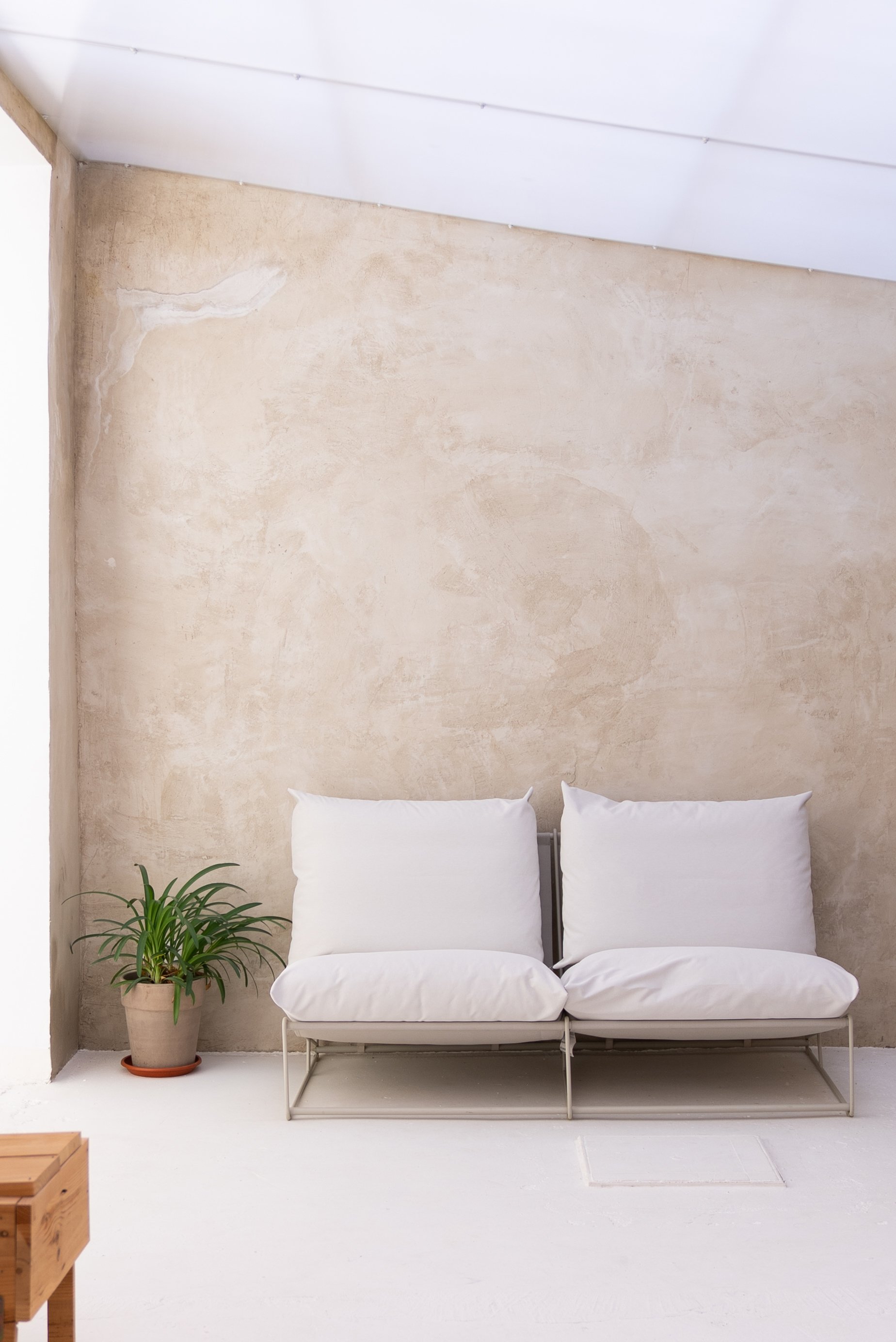 A minimalist indoor space with a beige wall, a white metal bench with white cushions, a green potted plant on the left, and a small wooden table in the left foreground.