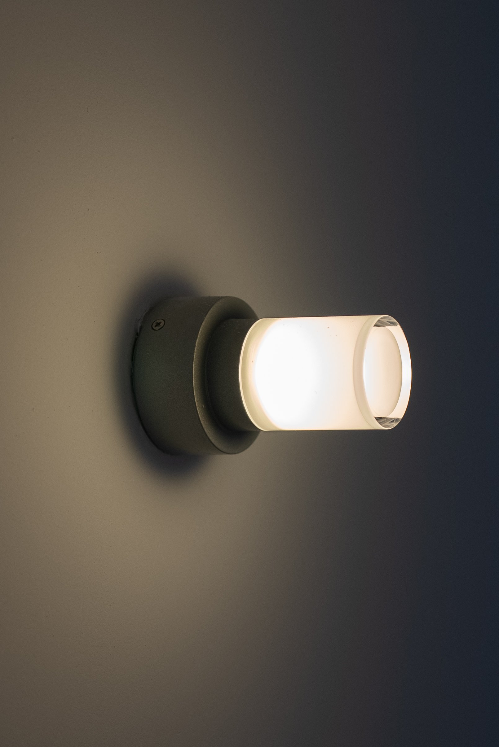 A modern wall-mounted light fixture with a cylindrical frosted glass shade on a nearby wall, emitting a soft white light.