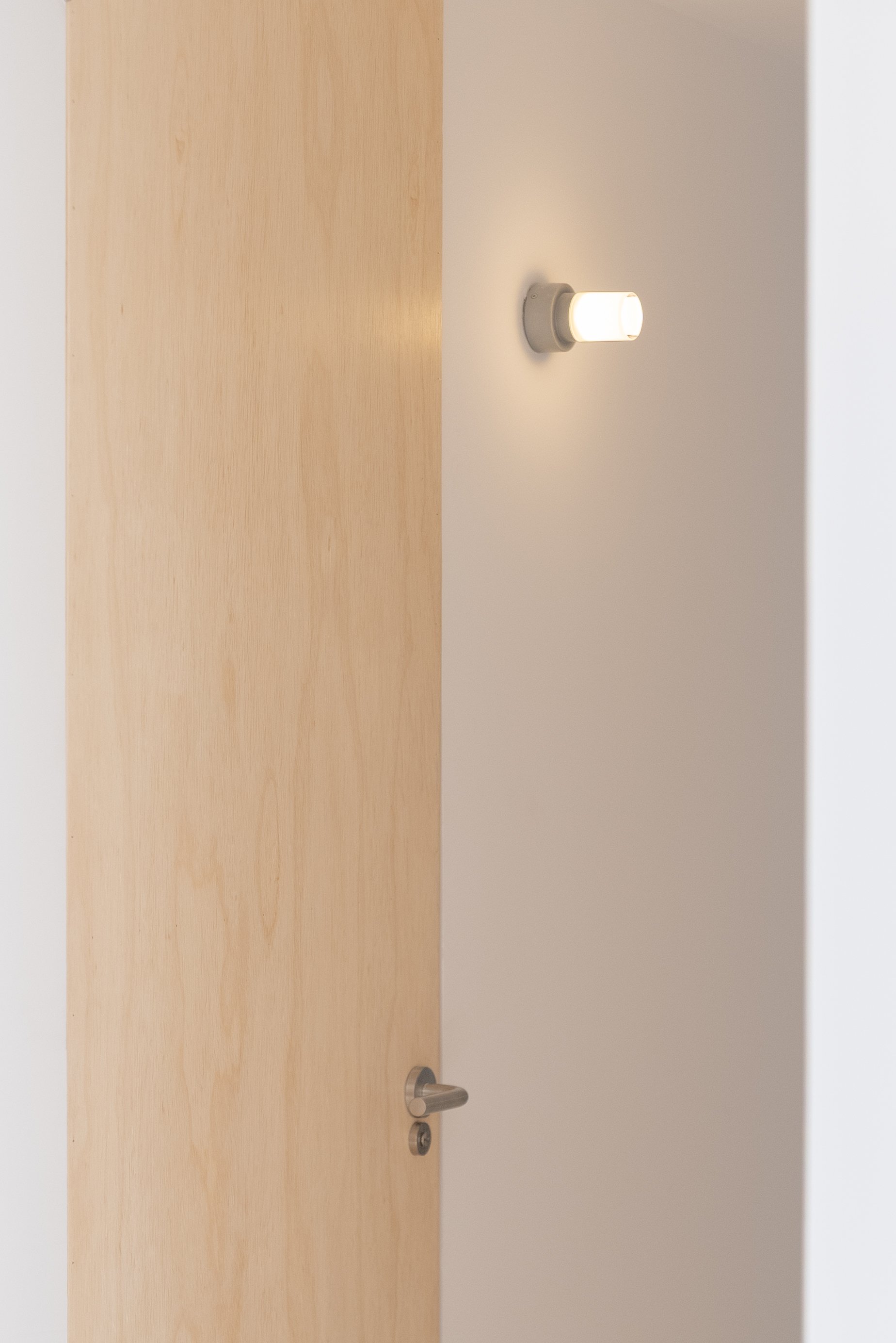 Partially opened wooden door with a metallic door handle, showing a small glimpse of the interior and a wall-mounted light fixture nearby.