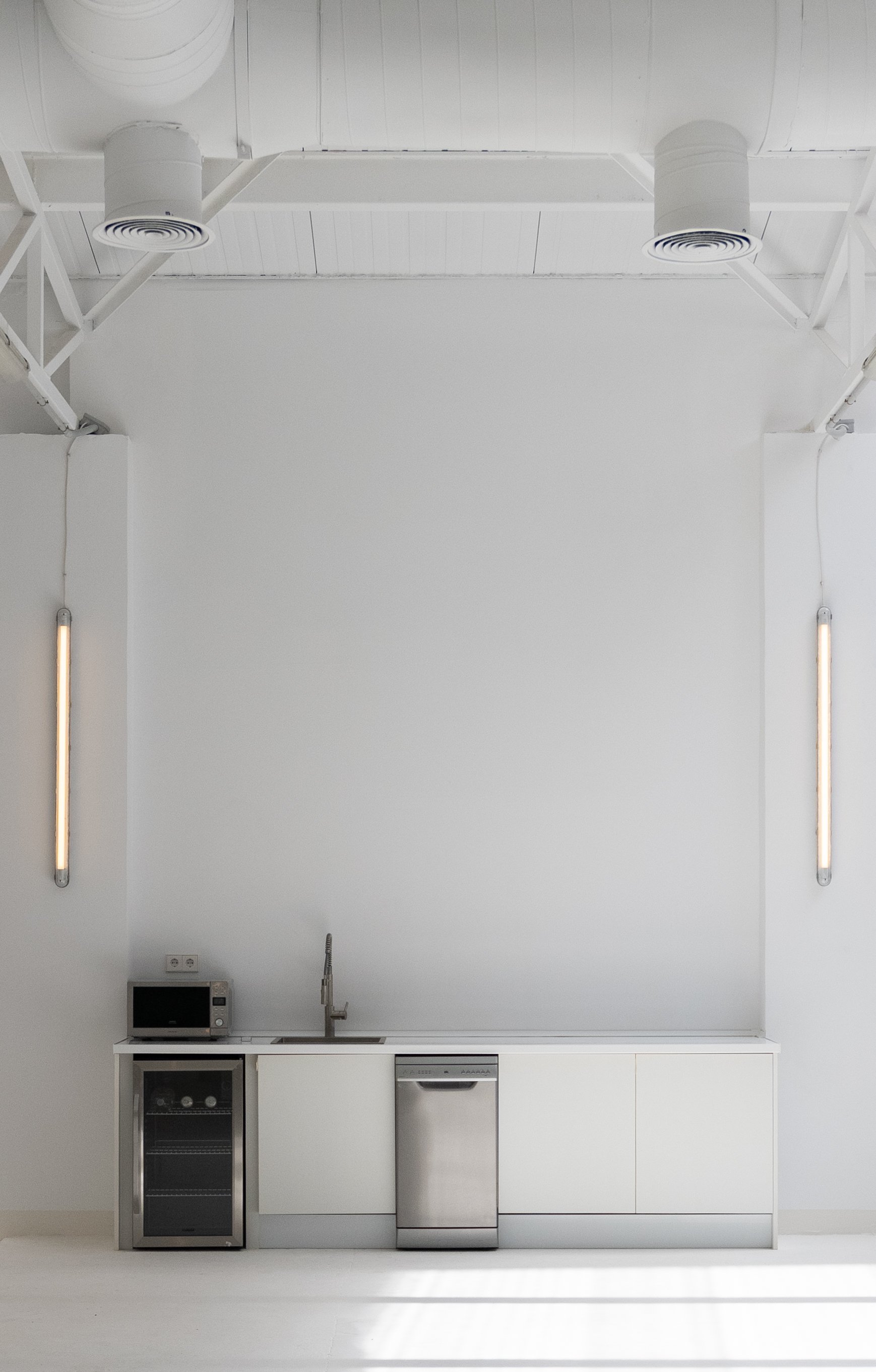 Minimalist kitchen with white cabinets, stainless steel appliances, microwave, small wine fridge, and dishwasher, set against a white wall in a bright room.