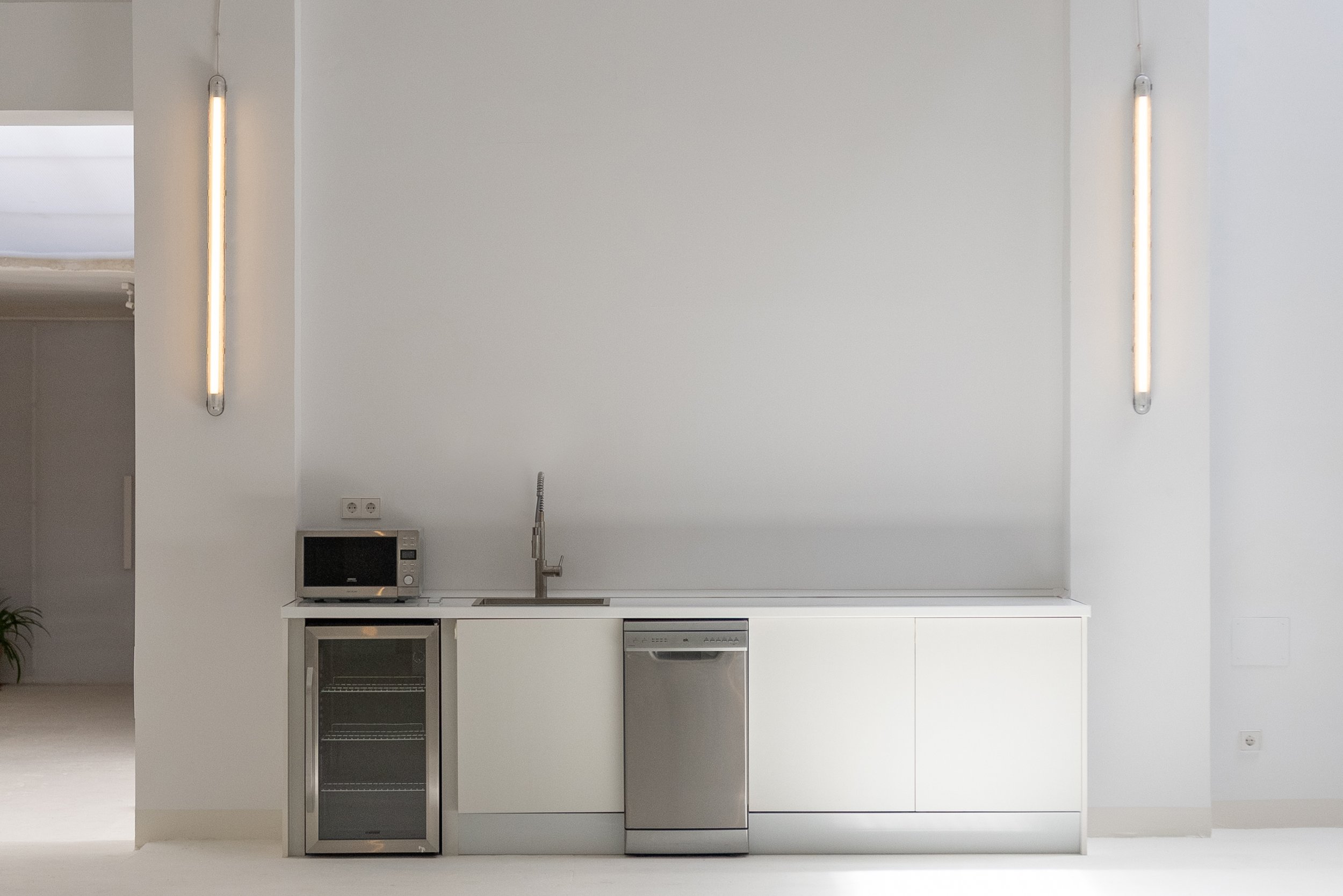 Minimalist kitchen with white cabinets, a small microwave, a wine fridge, and a dishwasher, with vertical wall lights on either side.