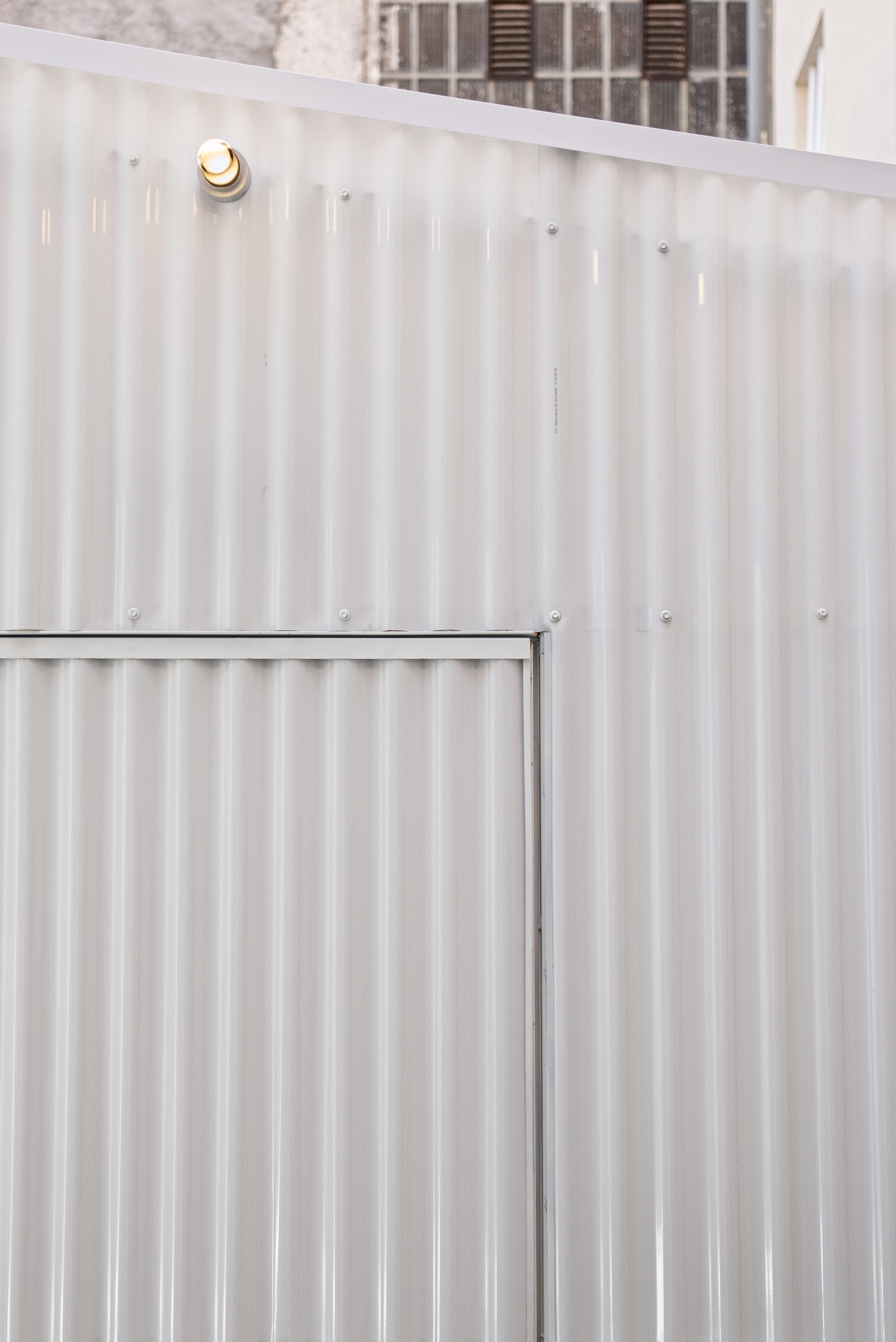 Close-up of a metallic, corrugated wall with a small round wall-mounted light at the top left and an angled roof above.
