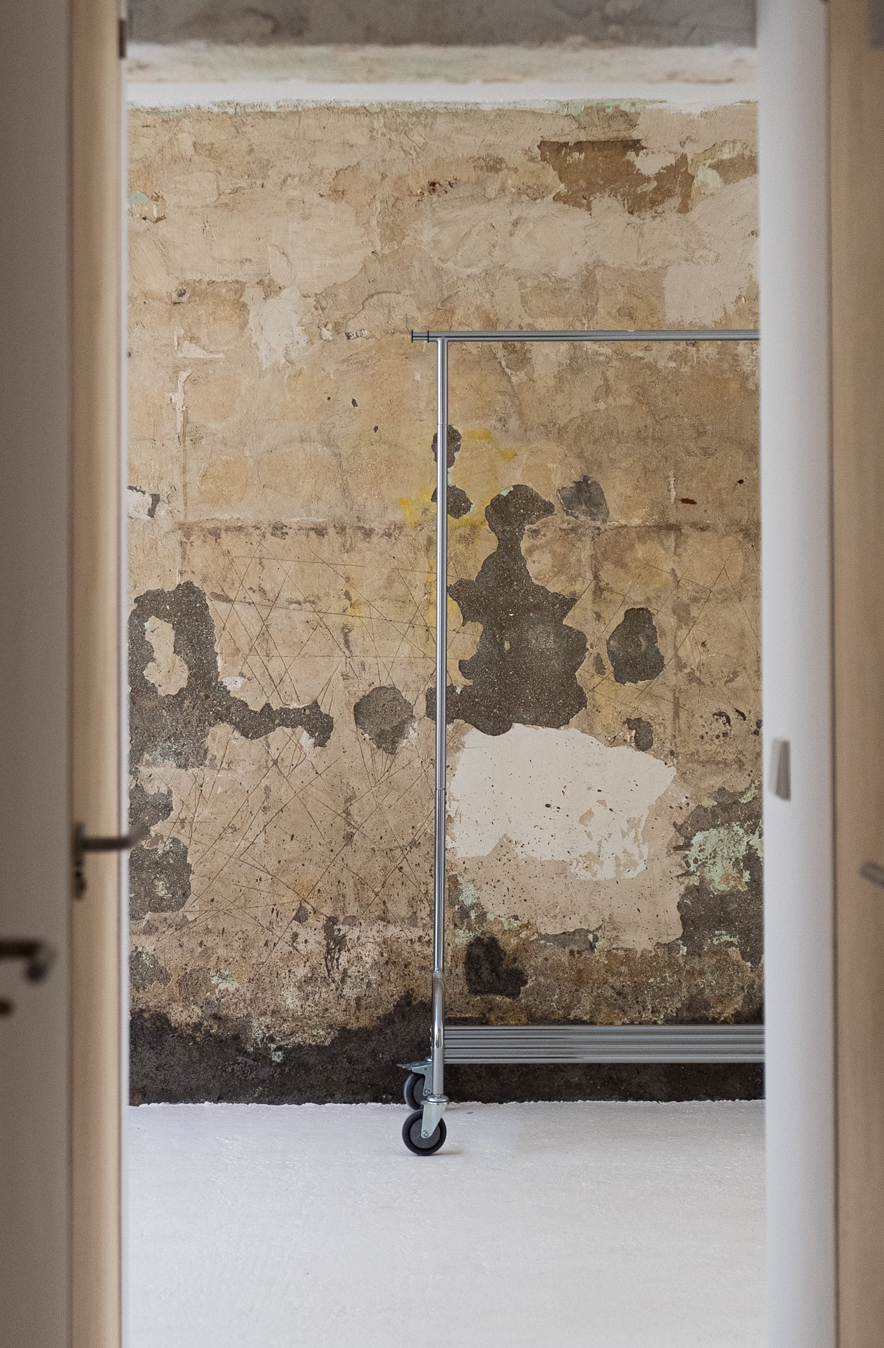 Empty room with a metal clothing rack on caster wheels against a weathered, peeling wall with patches of black and beige.