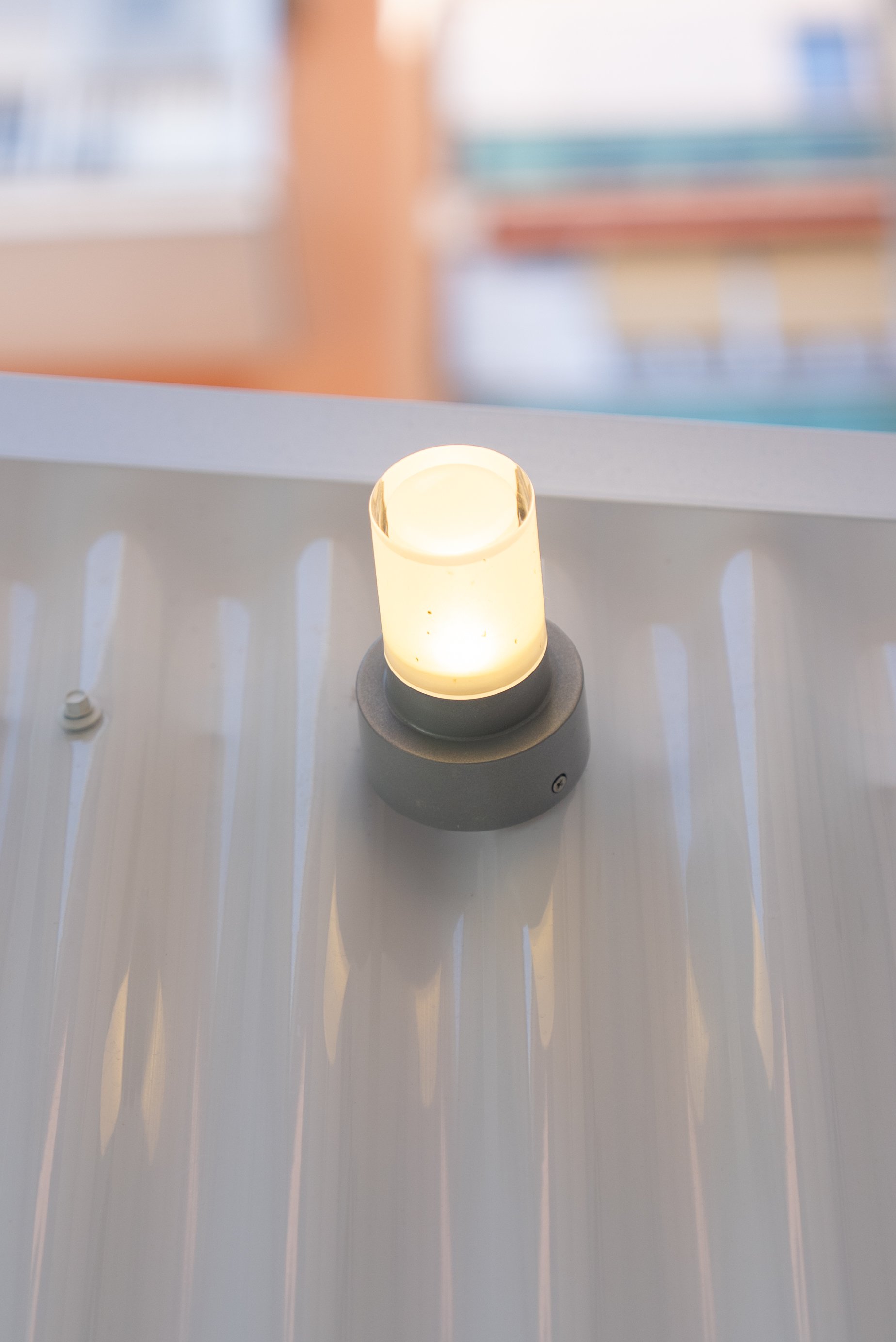 A glowing cylindrical wall-mounted light fixture on a shiny metallic surface with blurred background of buildings.