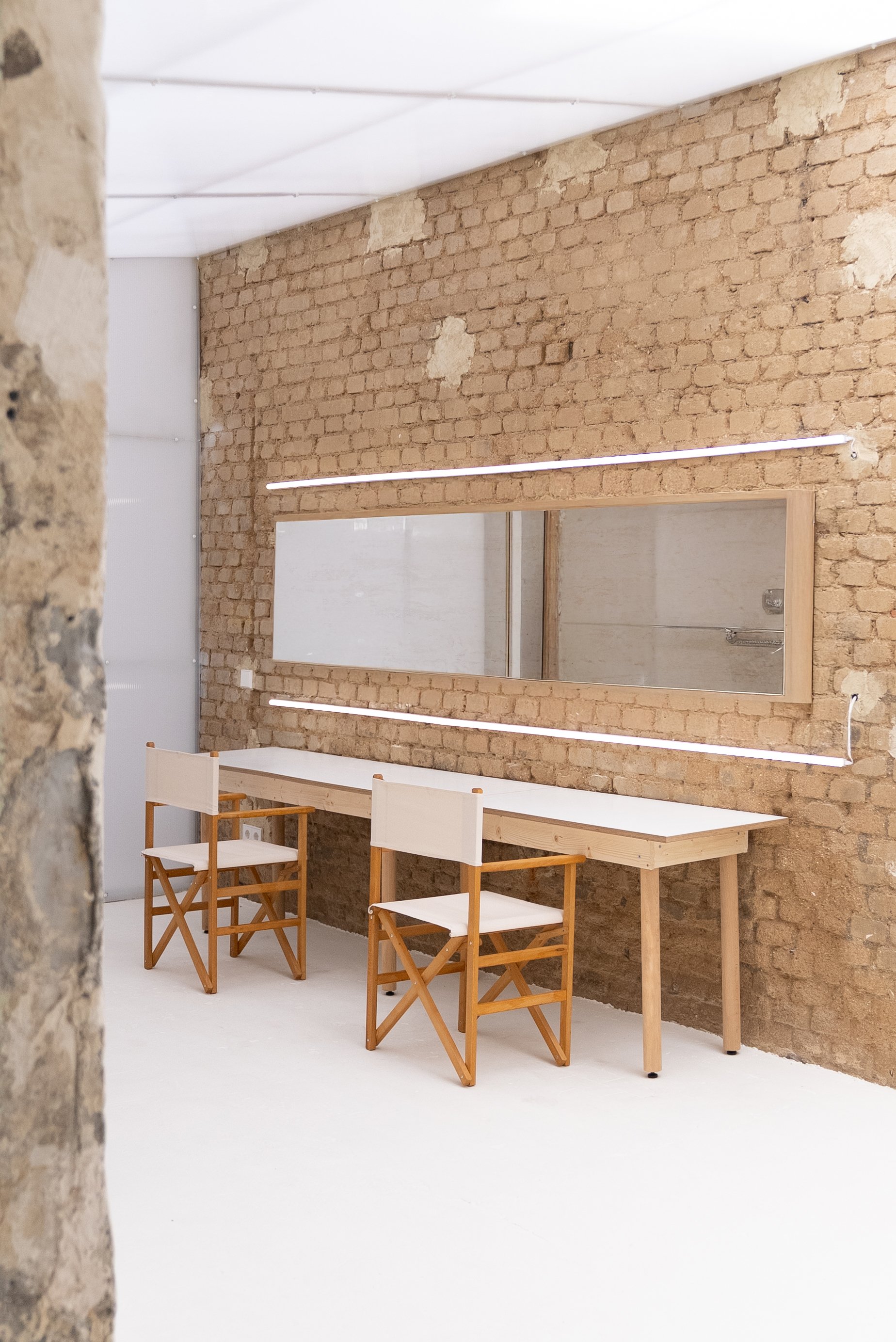 A minimalist workspace with a white table and two wooden chairs against a brick wall with a large mirror and linear LED lights.