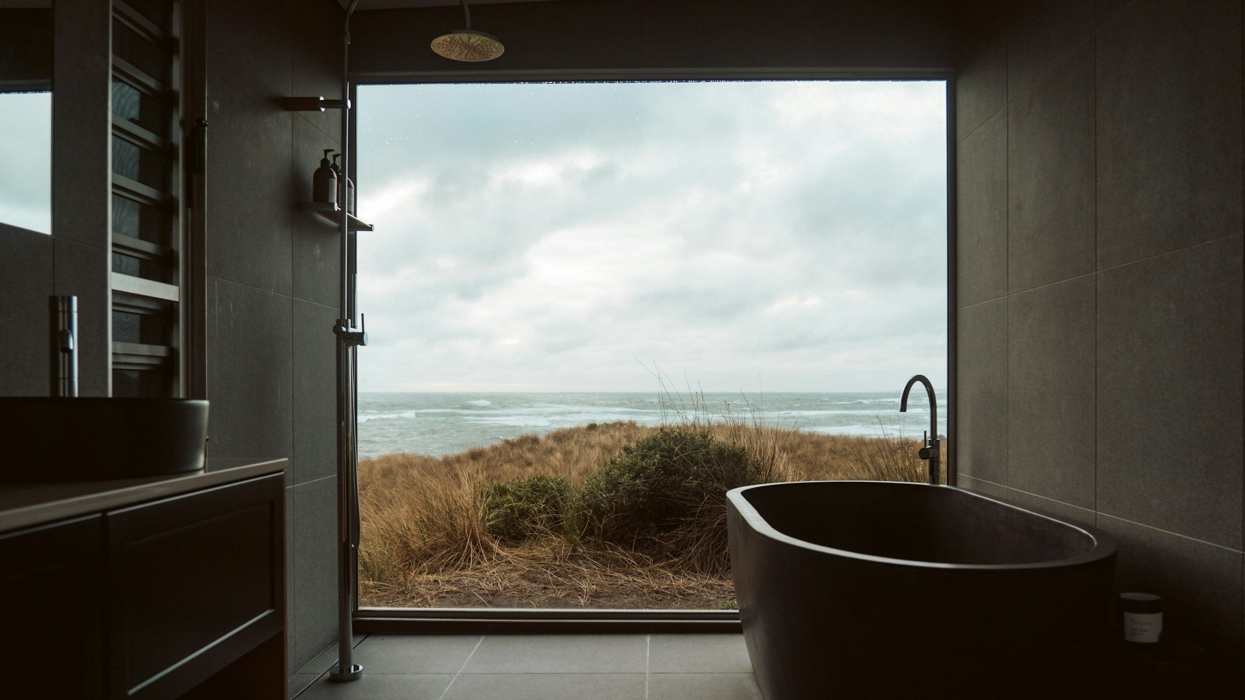 Modern bathroom with a large window overlooking a beach and ocean, featuring a black freestanding bathtub, dark tiled walls, and fixtures.