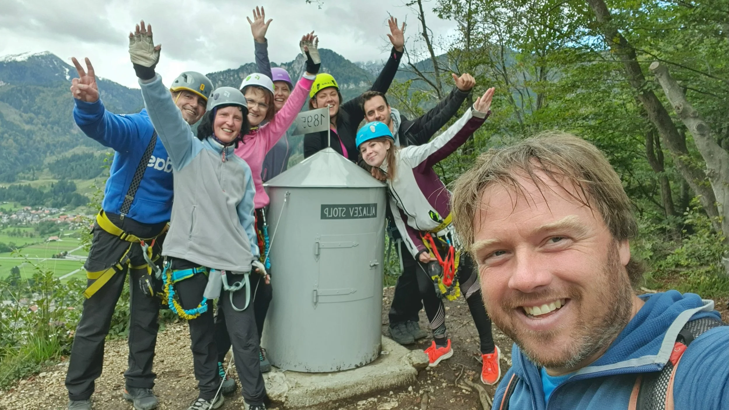 Group of seven climbers in outdoor gear celebrating at a mountain peak with a metal marker reading 'Alps' and a sign with coordinates, scenic mountains and greenery in the background.