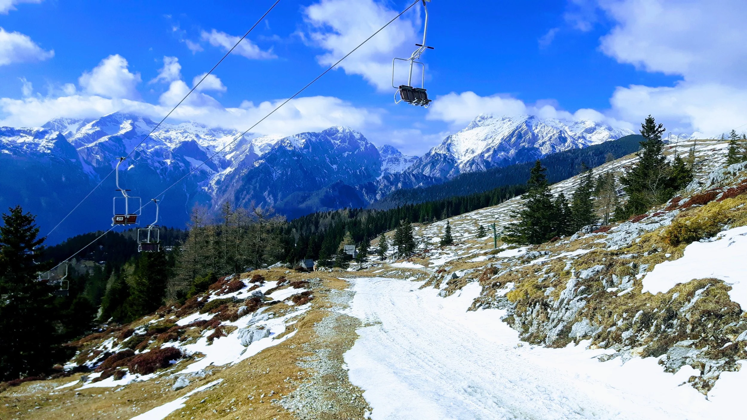 Snow-covered mountain landscape with pine trees, a dirt trail, and ski lifts under a partly cloudy sky.