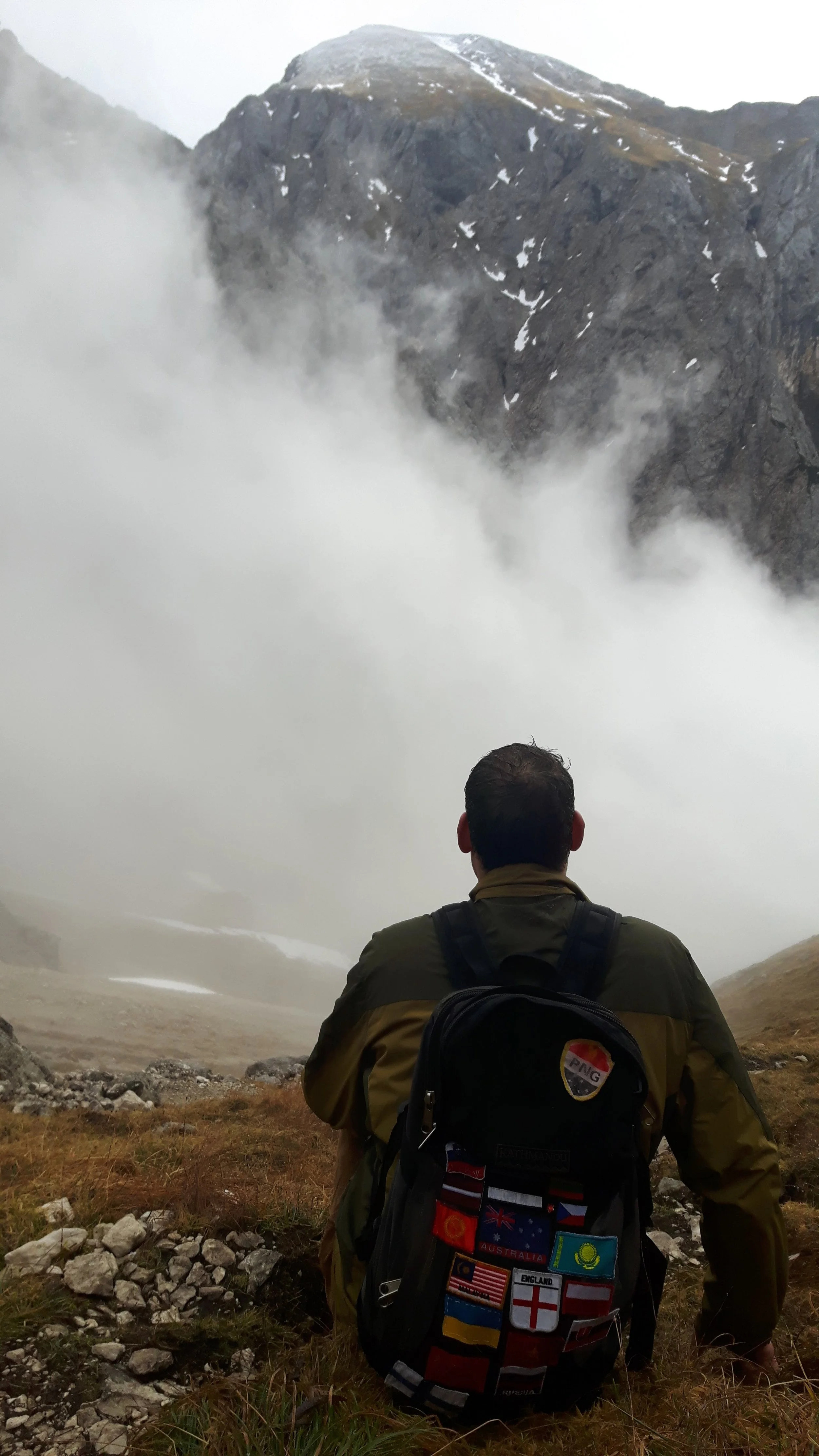 Back of a man sitting on rocky ground, wearing a backpack with various country flags, overlooking a foggy mountainous landscape with snow patches.