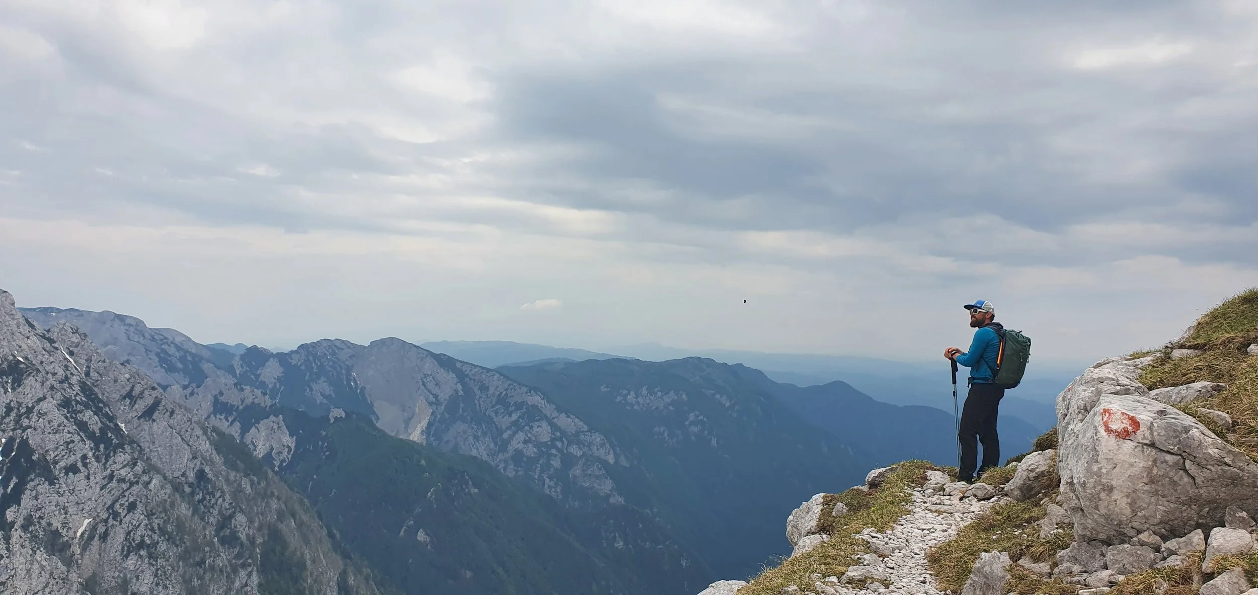 Hiker with a backpack and trekking poles standing on a rocky mountain trail overlooking a mountain range under a cloudy sky.