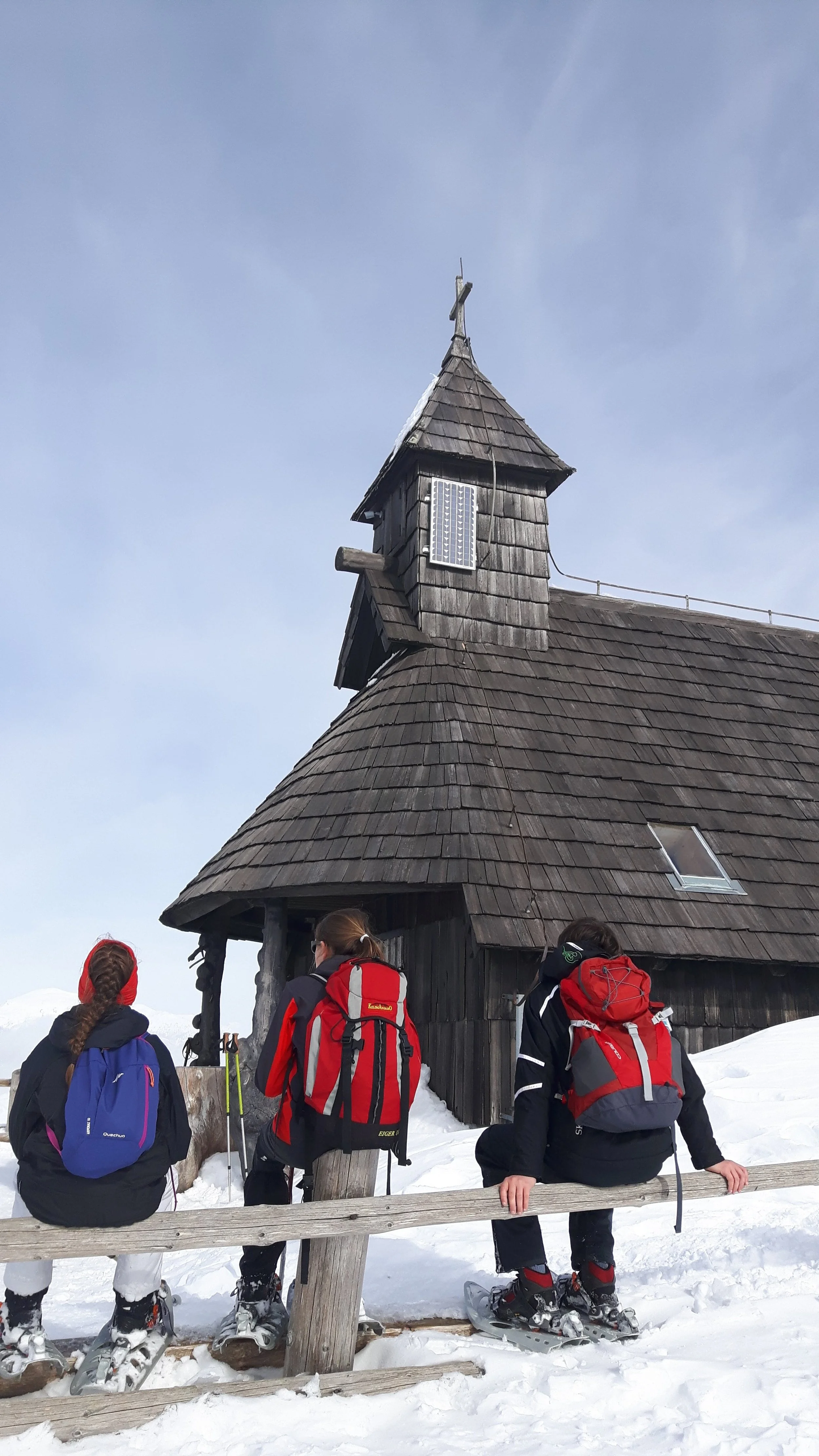 Three people with backpacks and snowshoes sitting on a wooden fence in front of a small black wooden church with a shingled roof, in a snowy landscape under a cloudy sky.