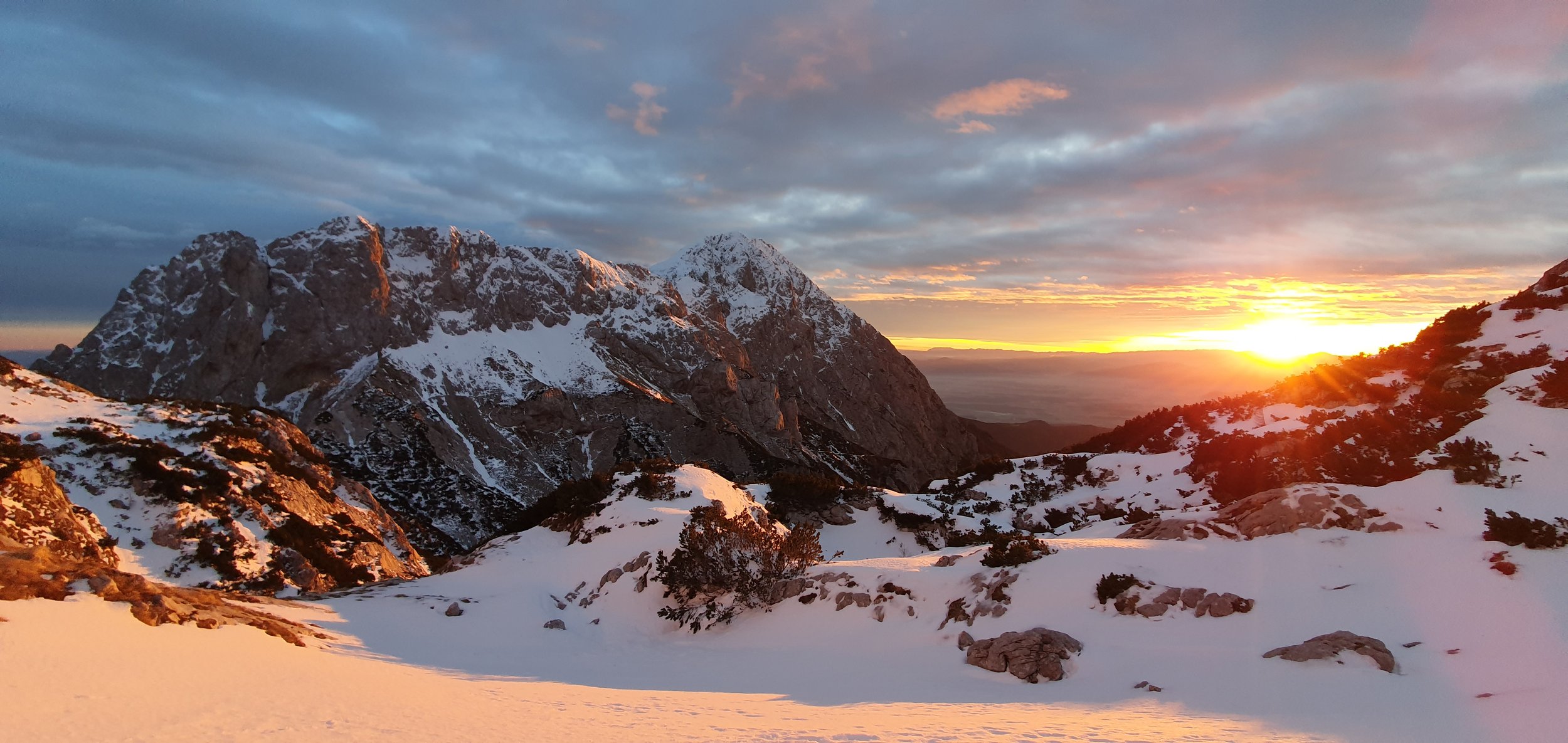 Snow-covered mountains during sunset with a cloudy sky, rocky terrain, and sparse bushes in the foreground.
