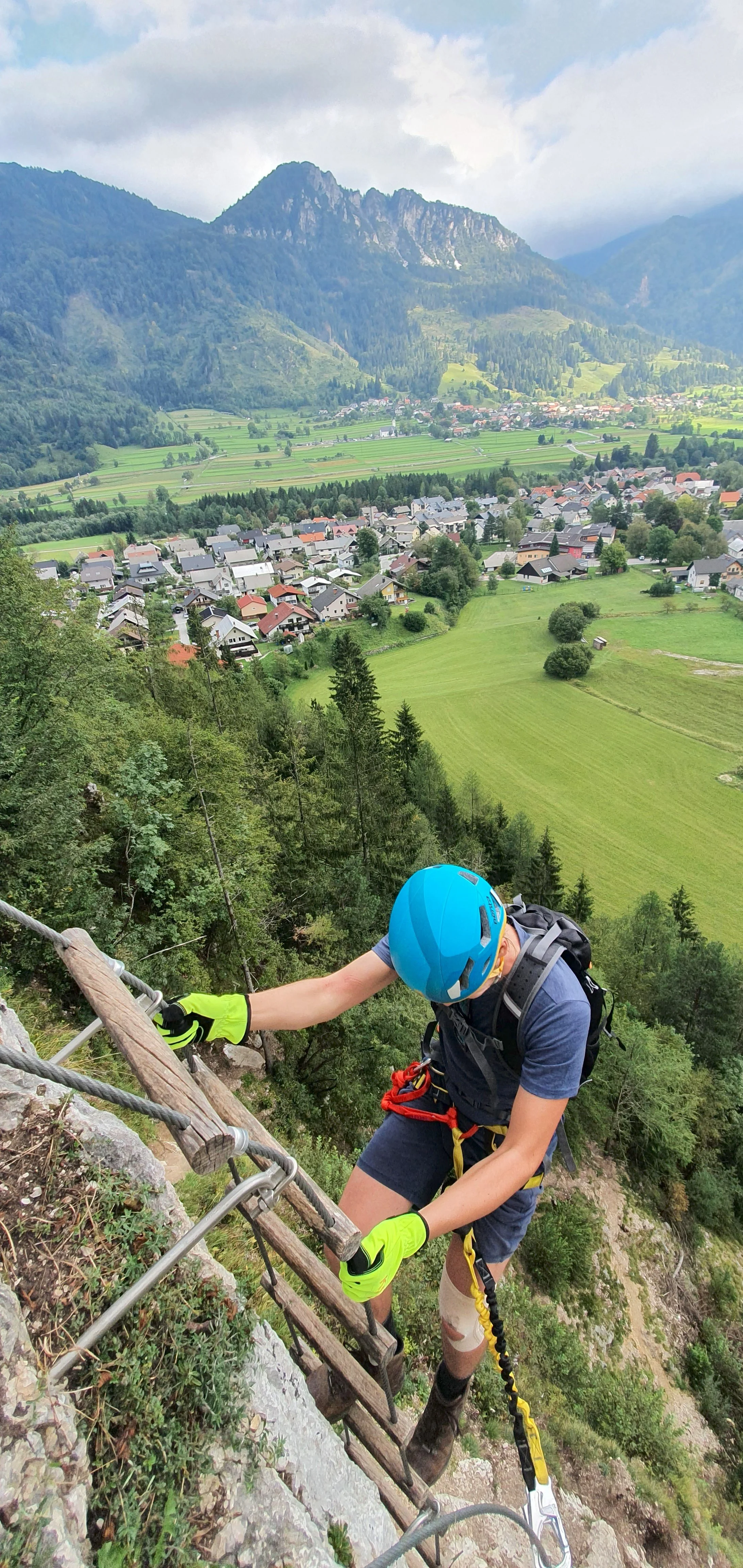 A person wearing a blue helmet and yellow gloves climbing a metal ladder on a mountainside, with a scenic view of a valley, green fields, a small village, and mountains in the background.