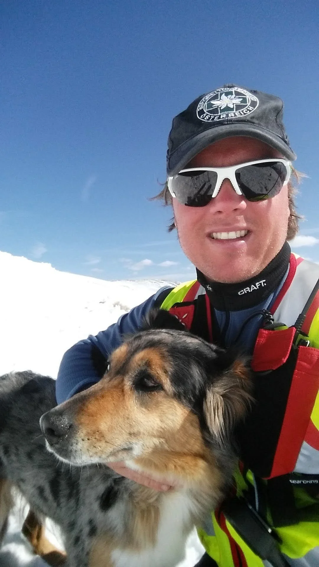 A man in sunglasses, a black cap, and a bright yellow and red jacket is outdoors in a snowy landscape with blue skies, holding a dog with black, brown, and white fur.