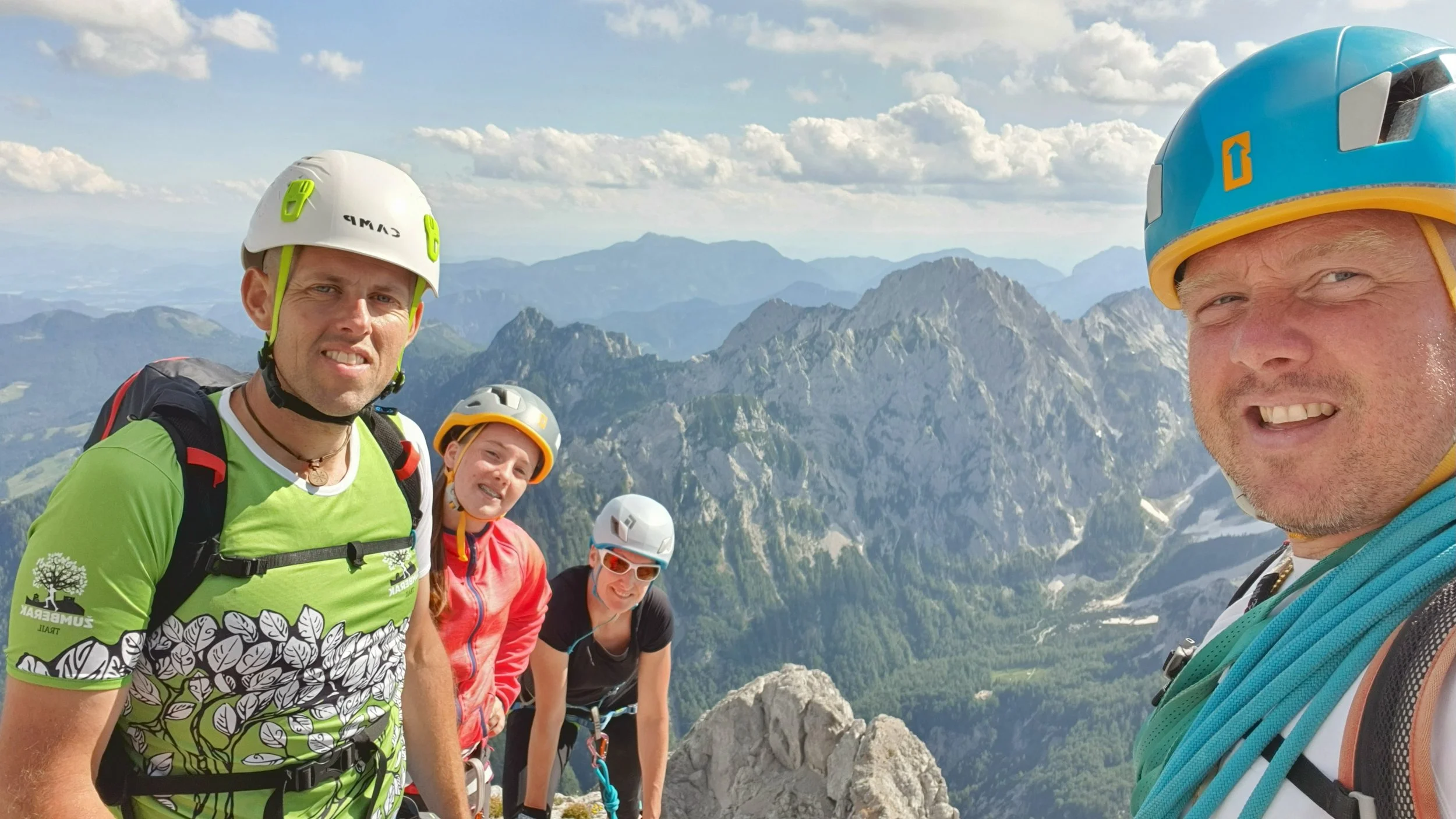 Four climbers wearing helmets and harnesses on a mountain peak with a valley and mountain range in the background.