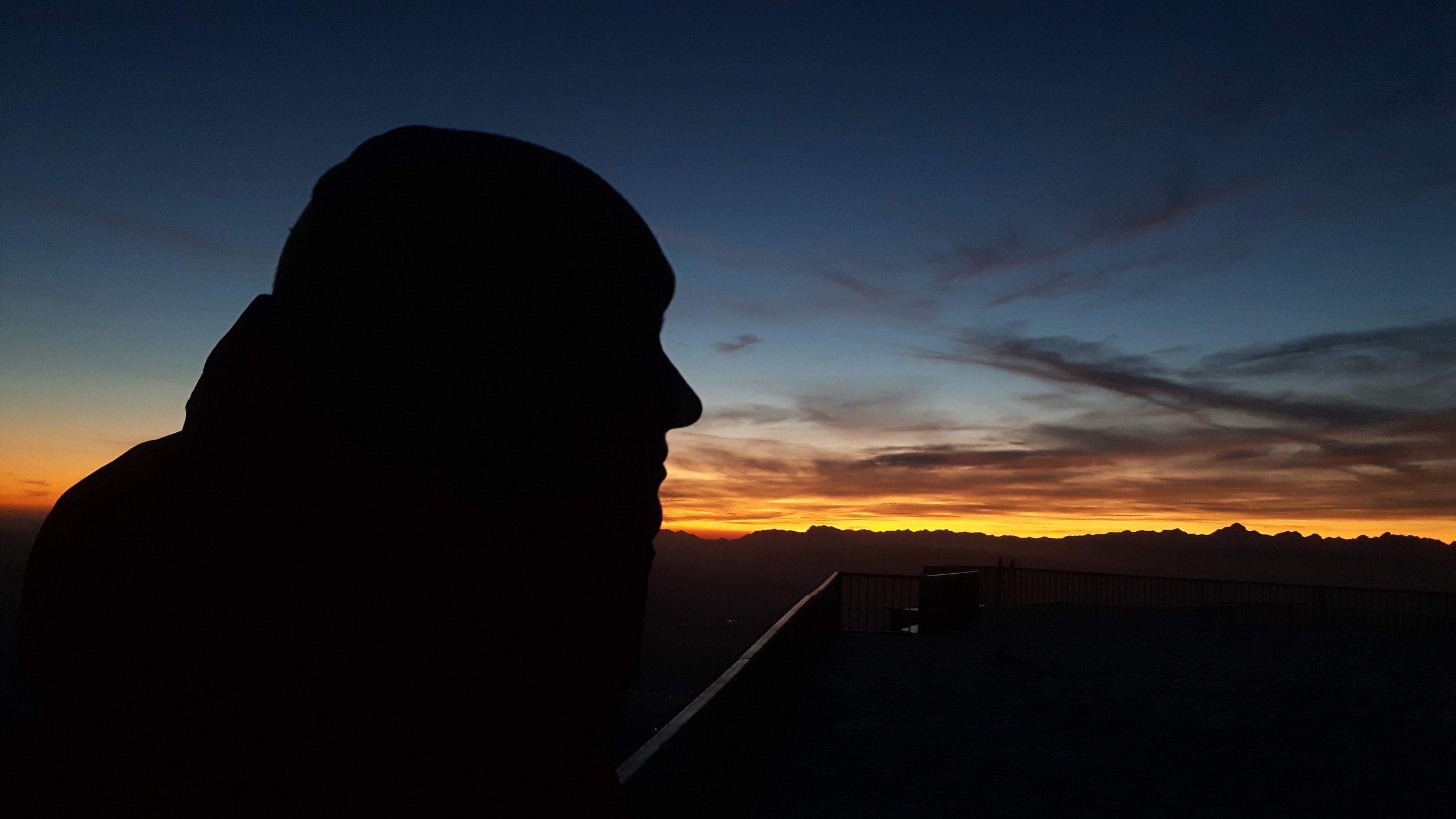 Silhouette of a person's profile against a colorful sunset sky with clouds and mountain ranges in the background.