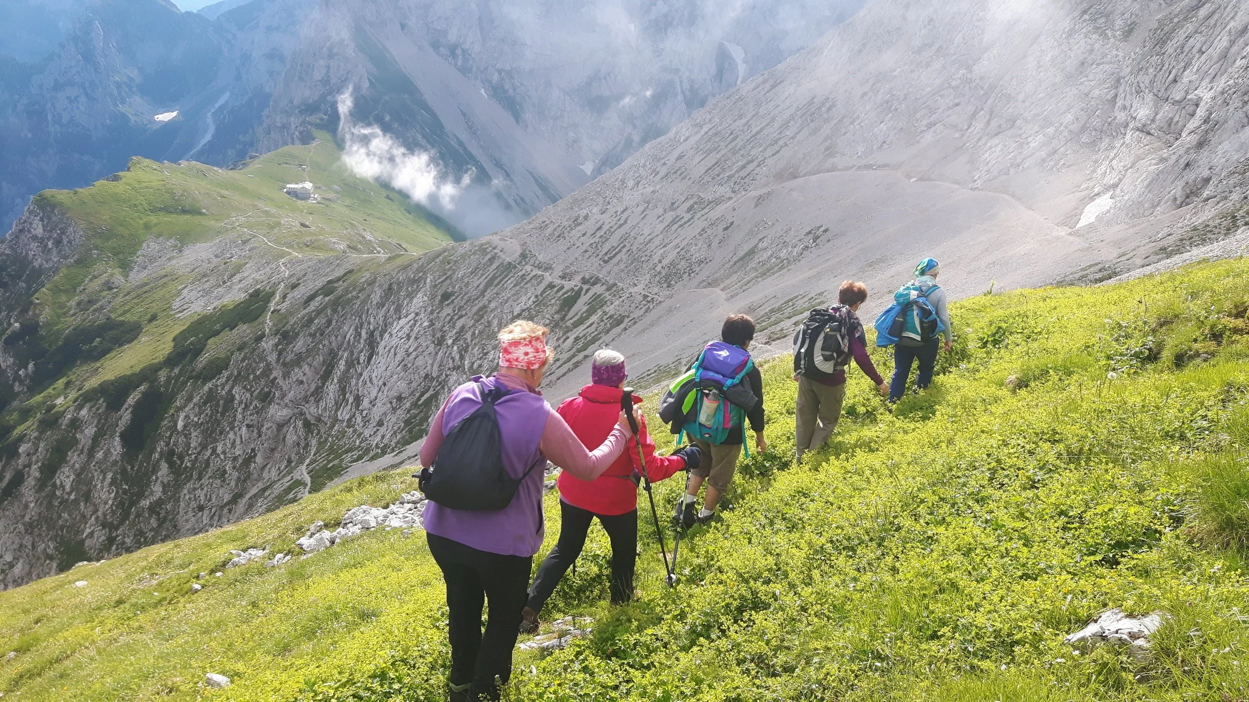 A group of six hikers, with backpacks and walking poles, trekking up a grassy mountain slope surrounded by rocky peaks and lush green valleys.